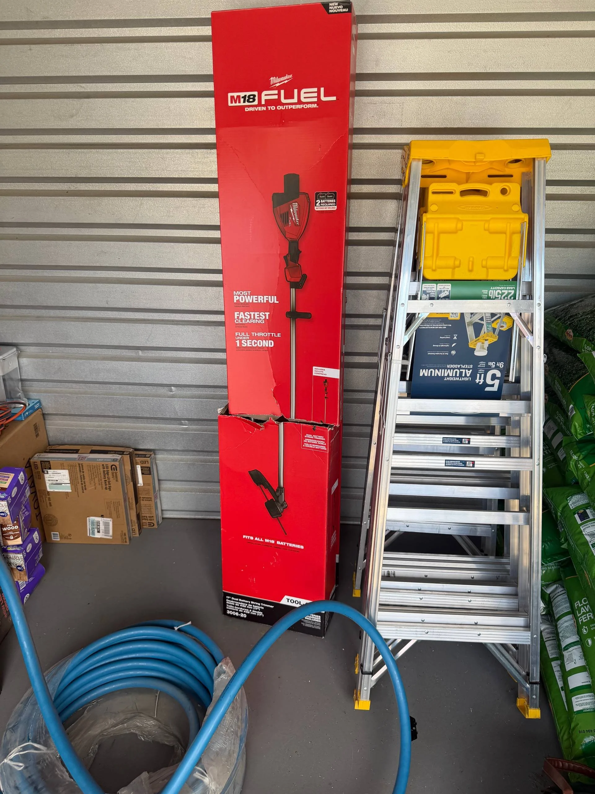 A red box with Milwaukee M18 Fuel cordless string trimmer packaging, a silver step ladder with yellow top, blue garden hose coiled on the floor, and green fertilizer bags stacked on the right.