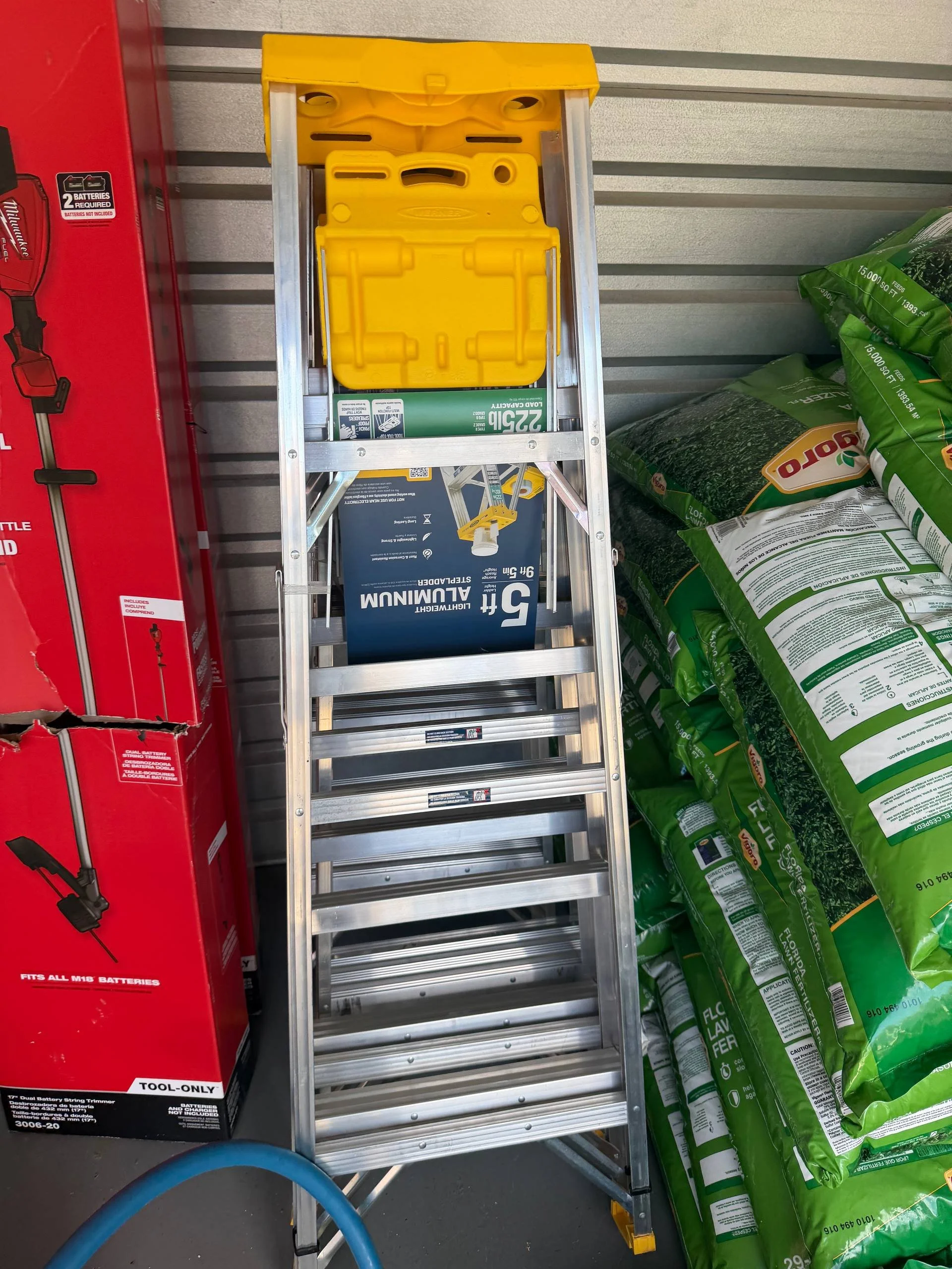 A step ladder with a yellow top tray, positioned between a red tool box and several large green bags of landscaping supplies in a hardware or garden store.