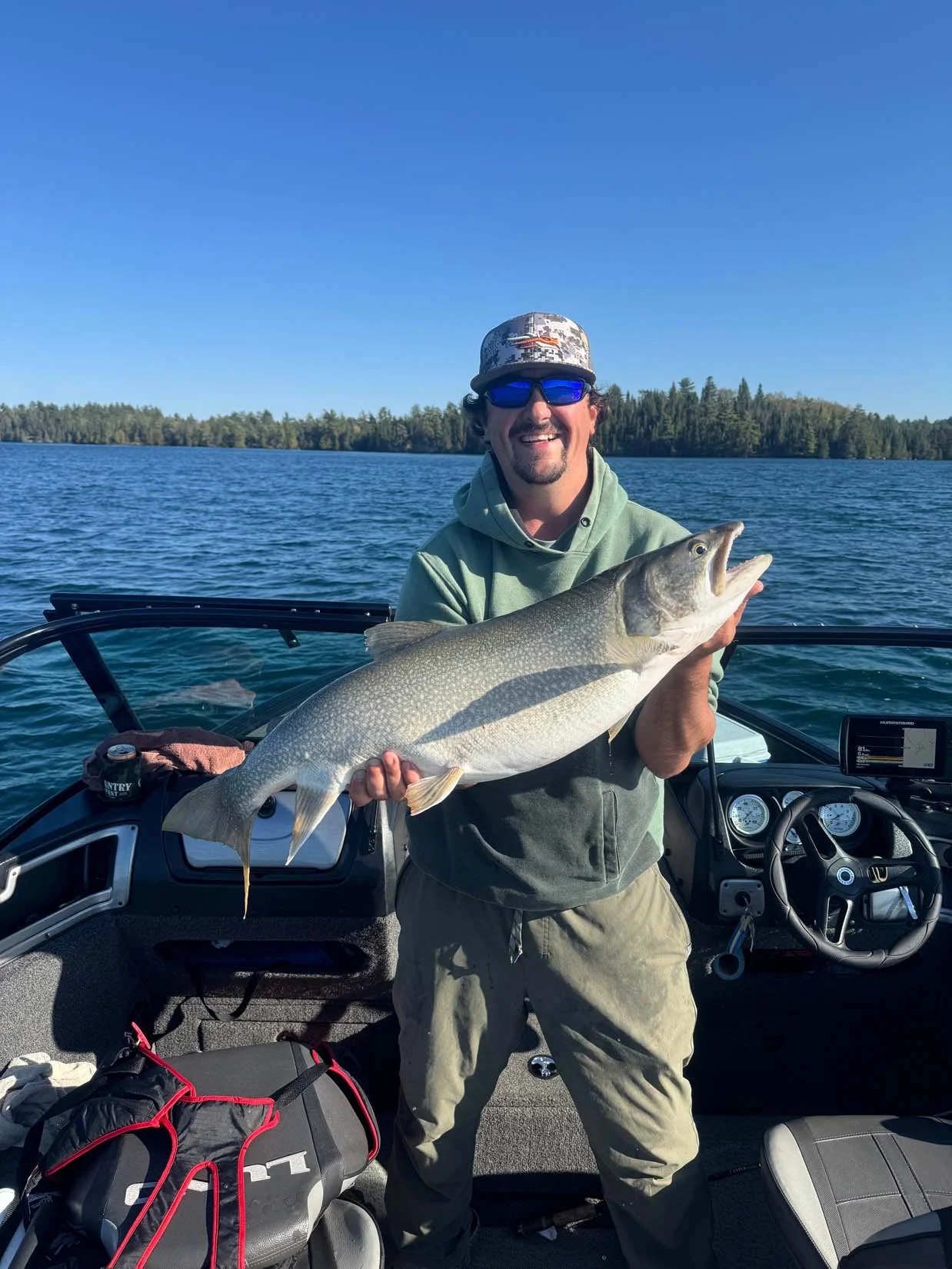 A man in a green hoodie and sunglasses smiling while holding a large fish on a boat in a lake with trees and a blue sky in the background.