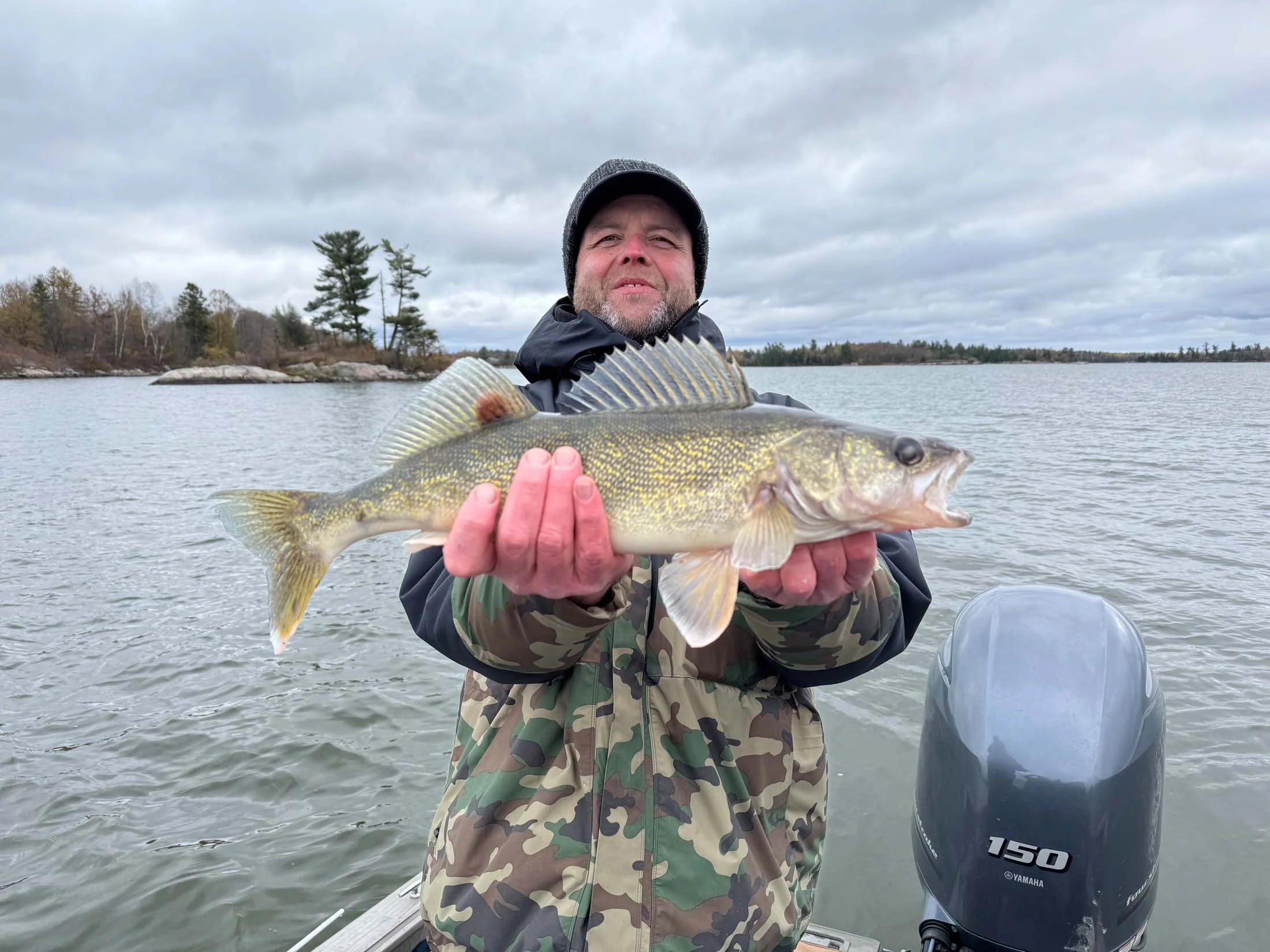 A man wearing camouflage jacket and gray beanie holding a large fish on a boat in a lake, with trees and cloudy sky in the background.
