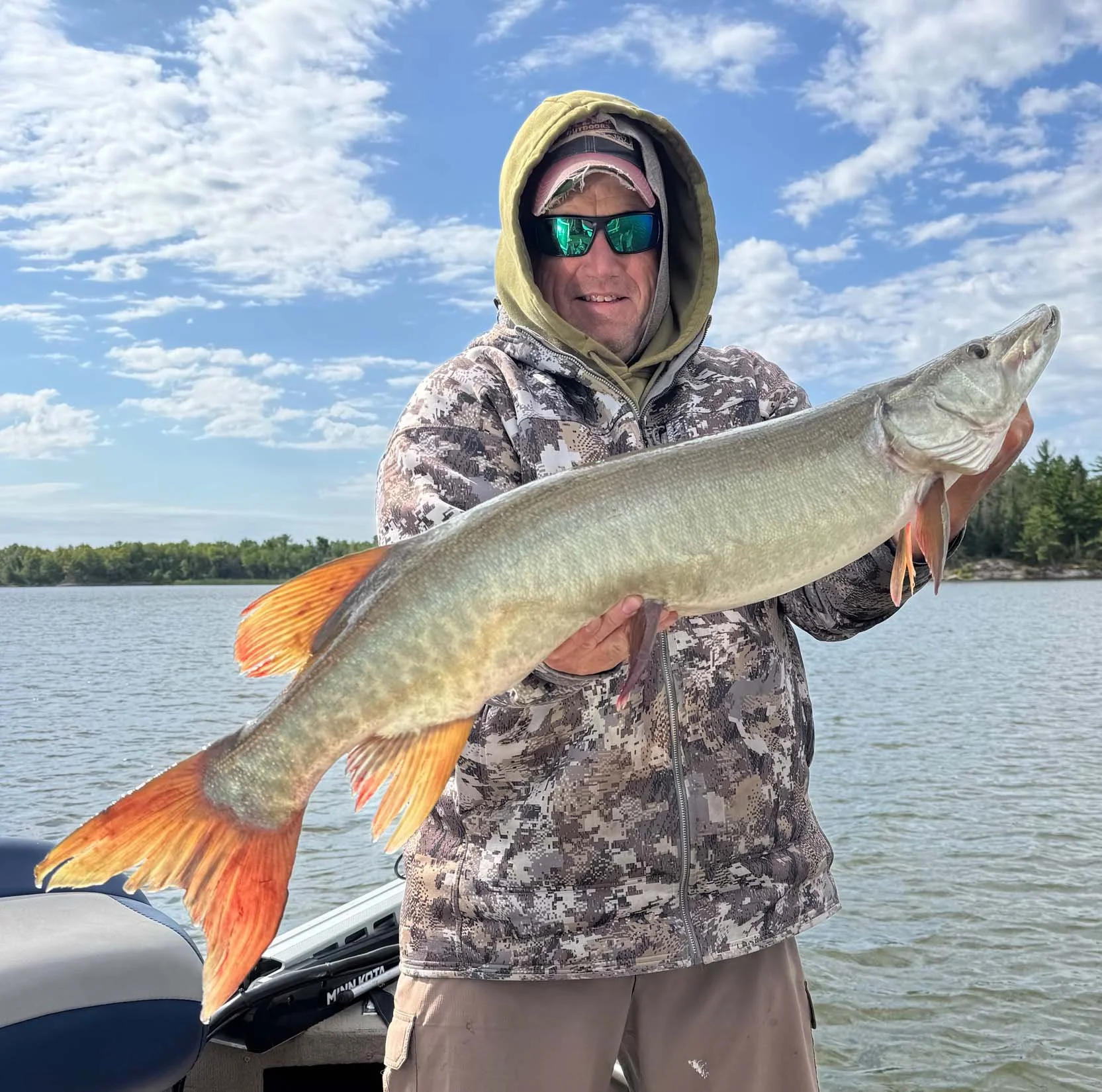 A man wearing sunglasses, a baseball cap, a hoodie, and a camouflage jacket near a body of water holding a large fish with orange fins.