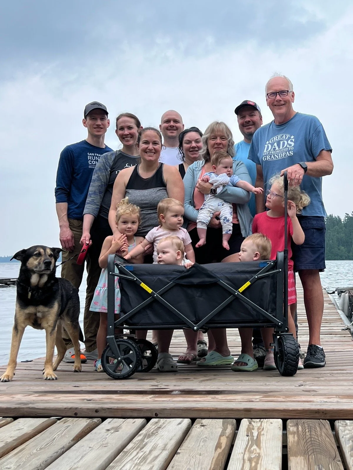 Family group photo on a wooden dock by a lake, including multiple children, adults, a dog, and a baby in a stroller, under cloudy sky.