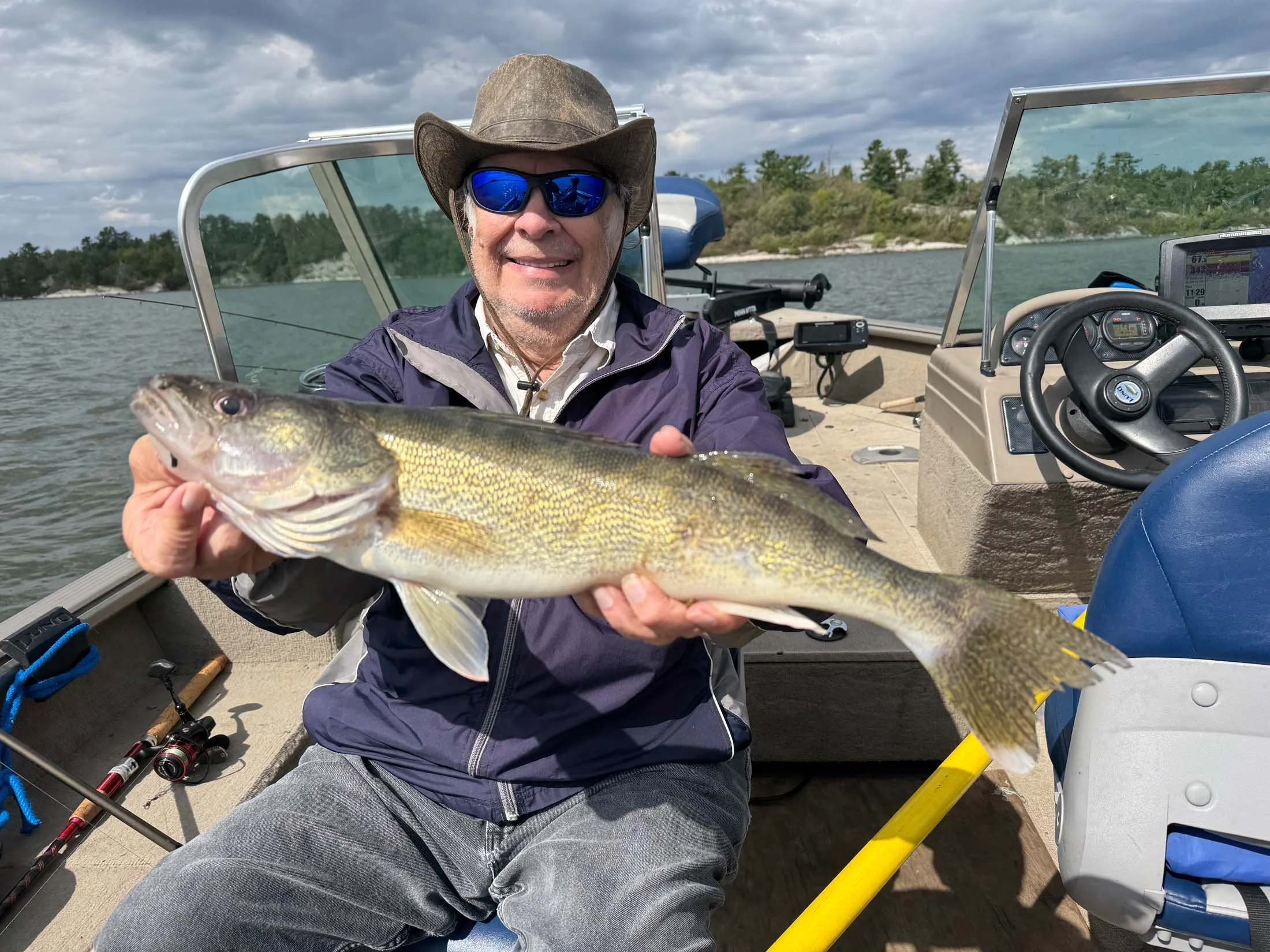 A man wearing sunglasses, a cowboy hat, and a purple jacket sitting on a boat holding a large fish, with fishing equipment visible around him and water and trees in the background.