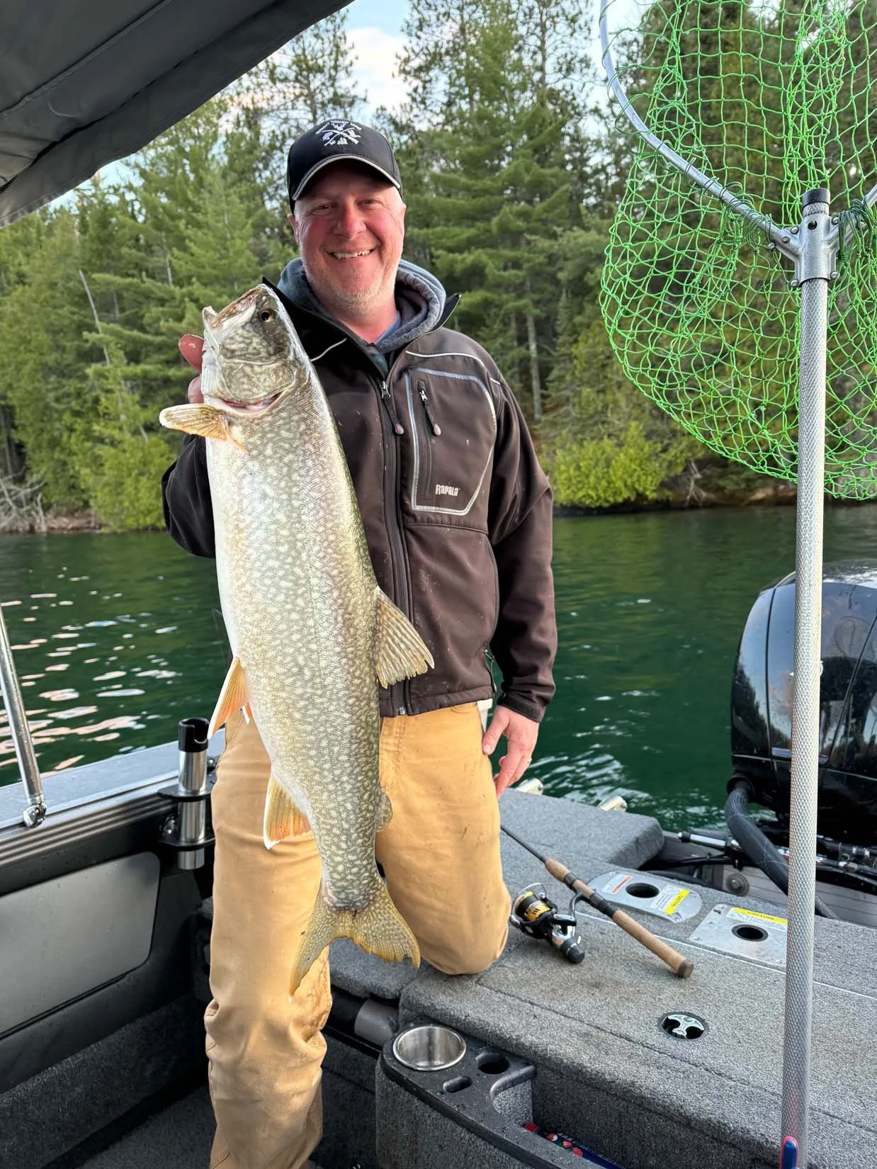 A man on a boat holding a large fish with trees in the background.