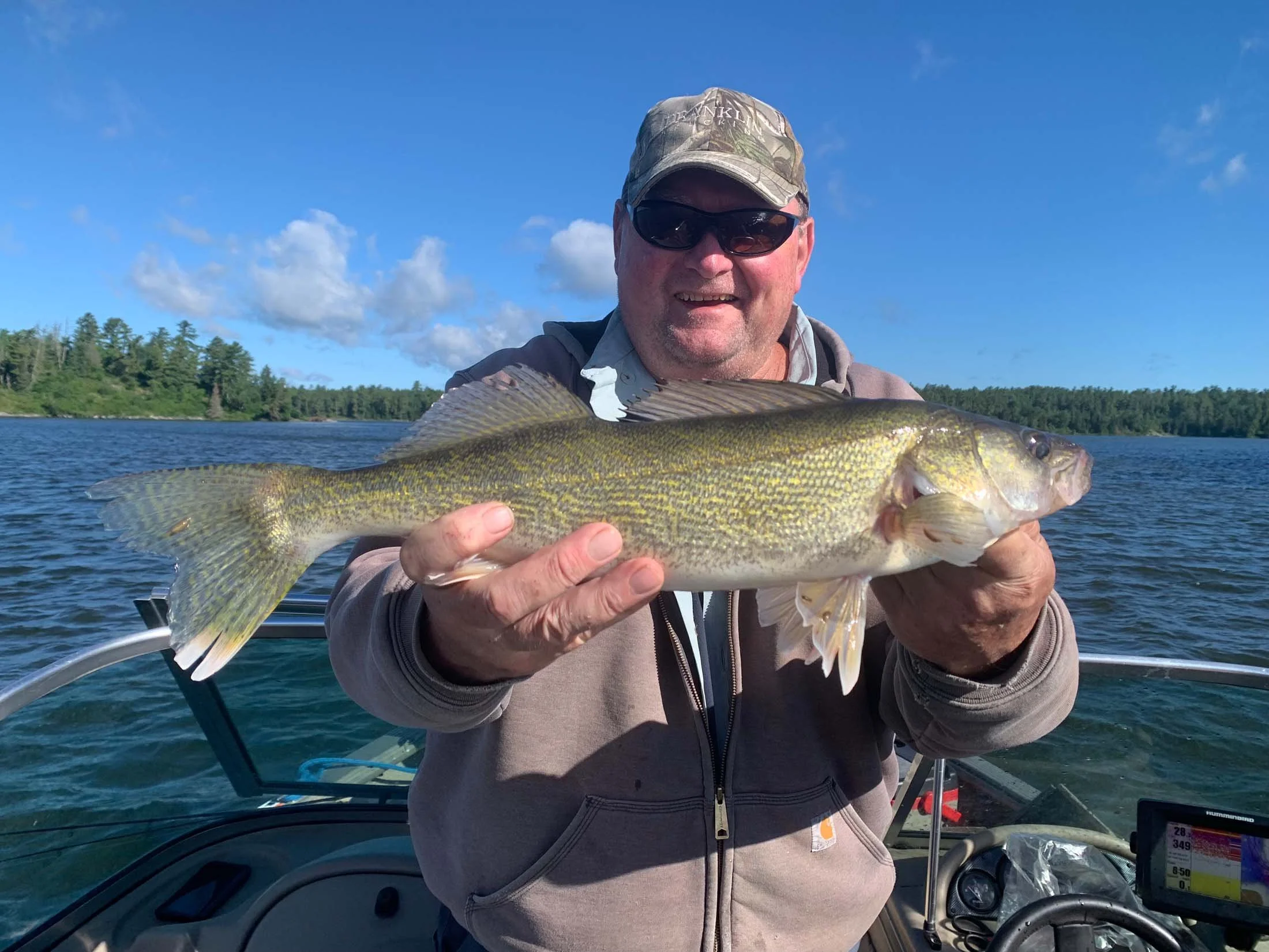 A man in sunglasses and a camouflage cap holding a large fish on a boat, with water and a tree-lined shoreline in the background under a partly cloudy sky.