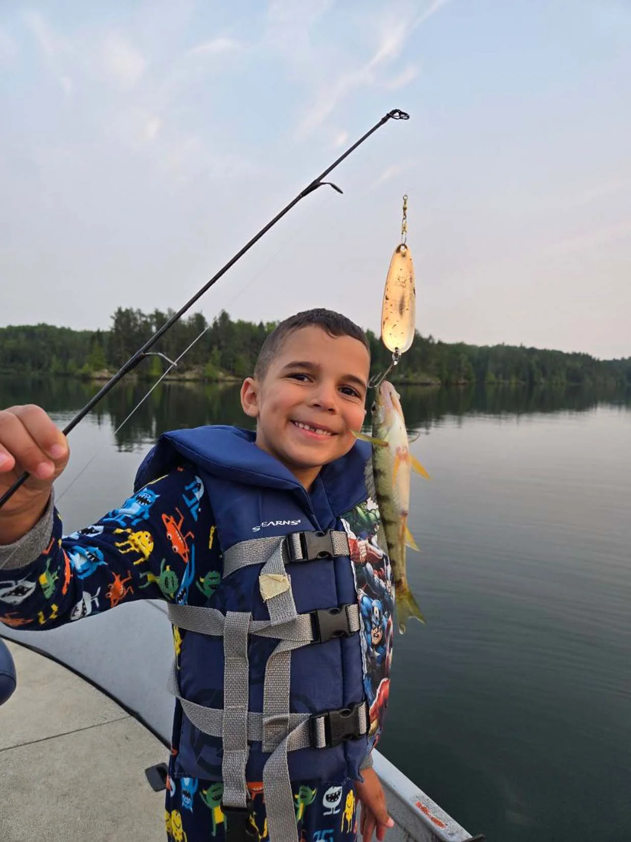 A boy smiling and holding up a small fish he caught on a fishing rod, standing on a dock near a calm lake with a forested shoreline in the background.