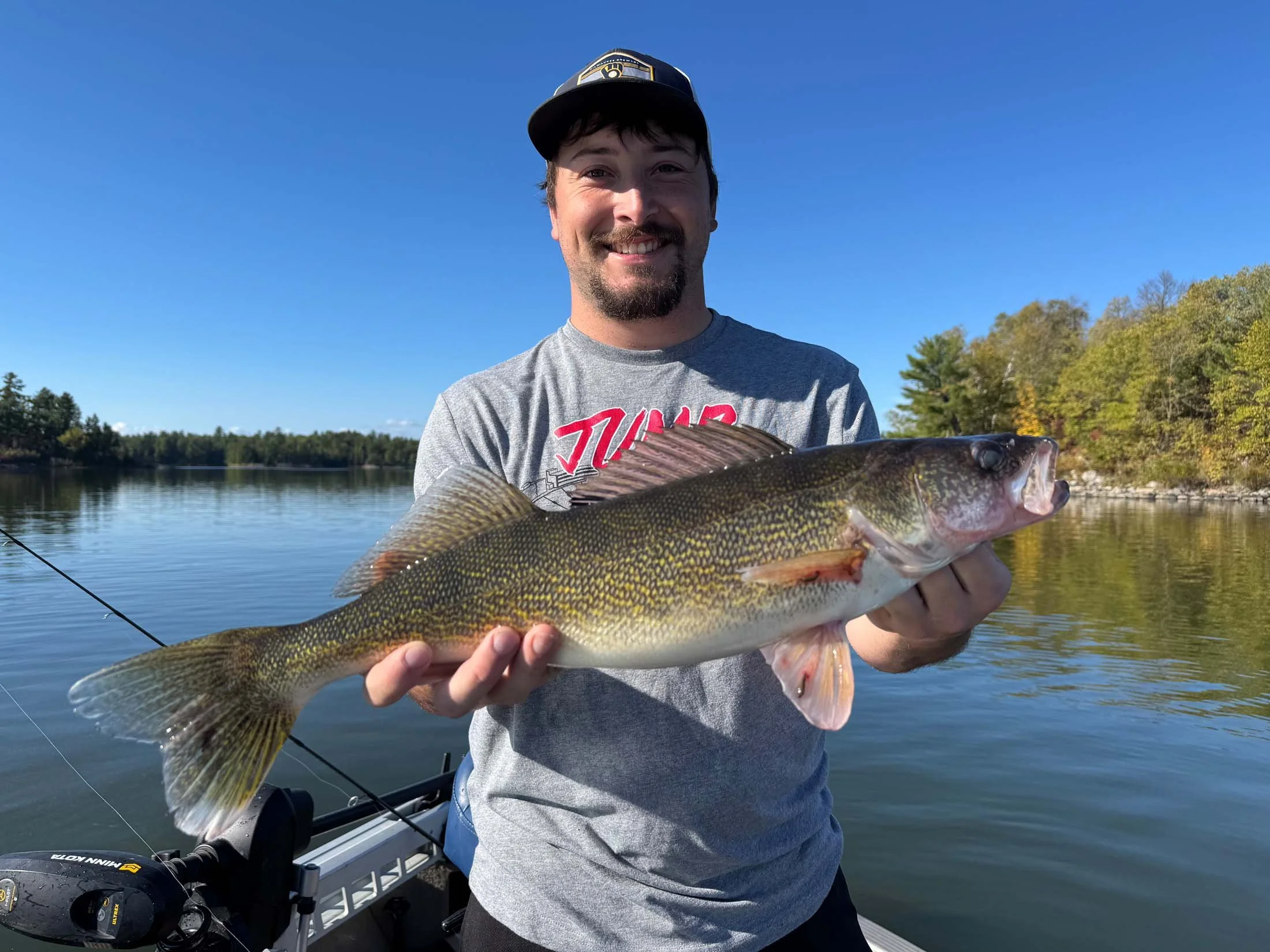 A man smiling and holding a large fish while standing on a boat with a lake and trees in the background.