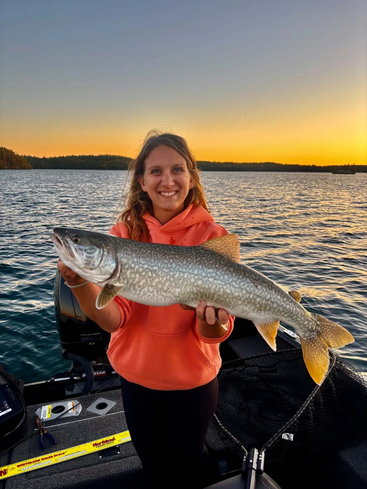 A woman smiling and holding a large fish on a boat during sunset over a body of water.