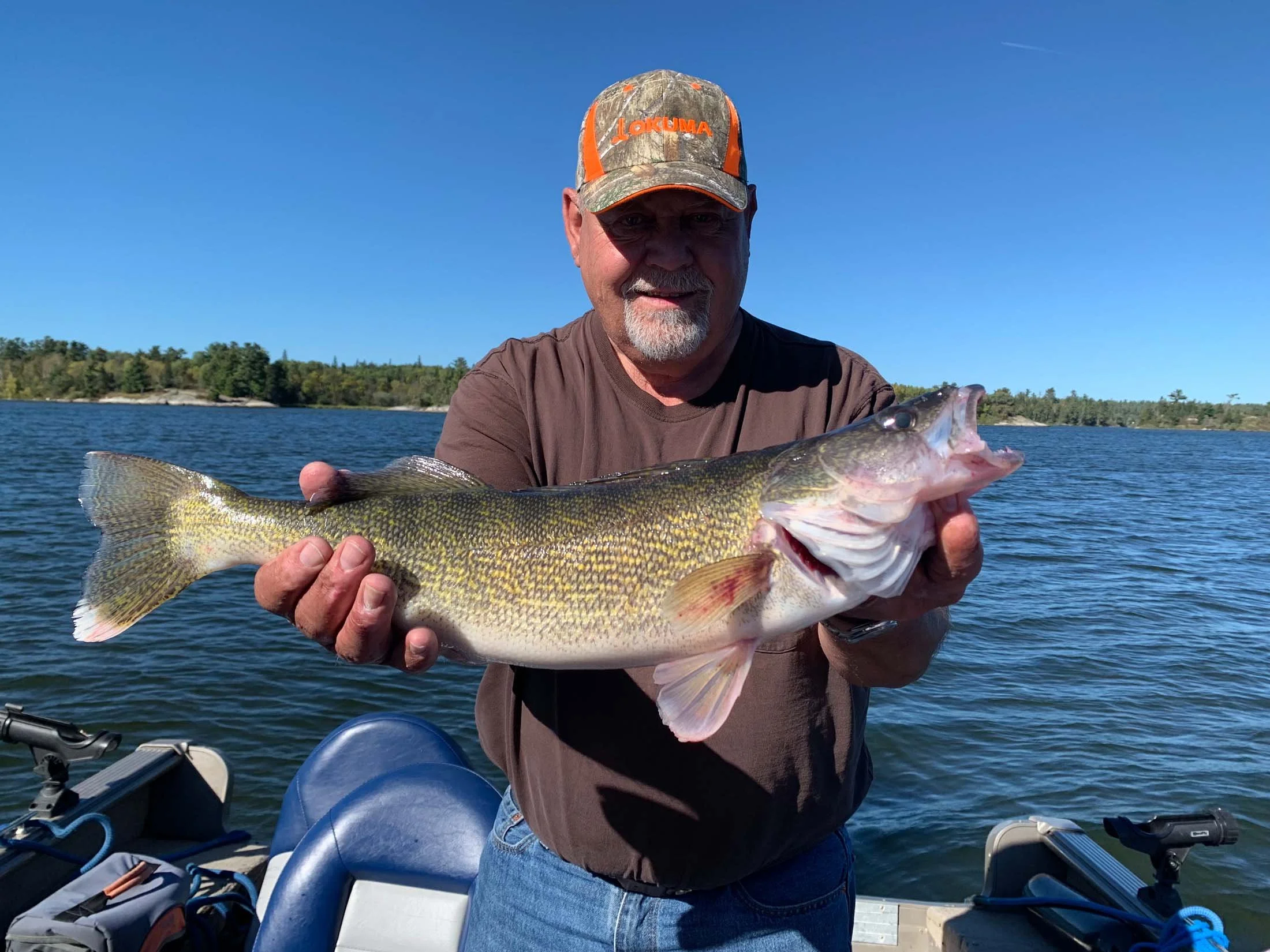 A man wearing a camouflage cap with orange lettering holding a large fish on a boat in a lake with a background of trees and blue sky.