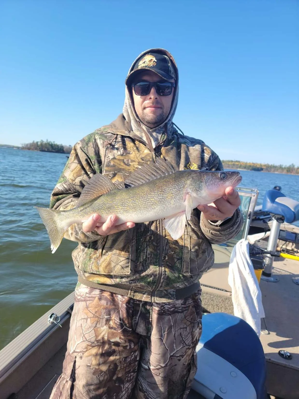 A man in camouflage clothes and black sunglasses on a boat holding a large fish he caught, with water and a distant shoreline in the background on a clear, sunny day.