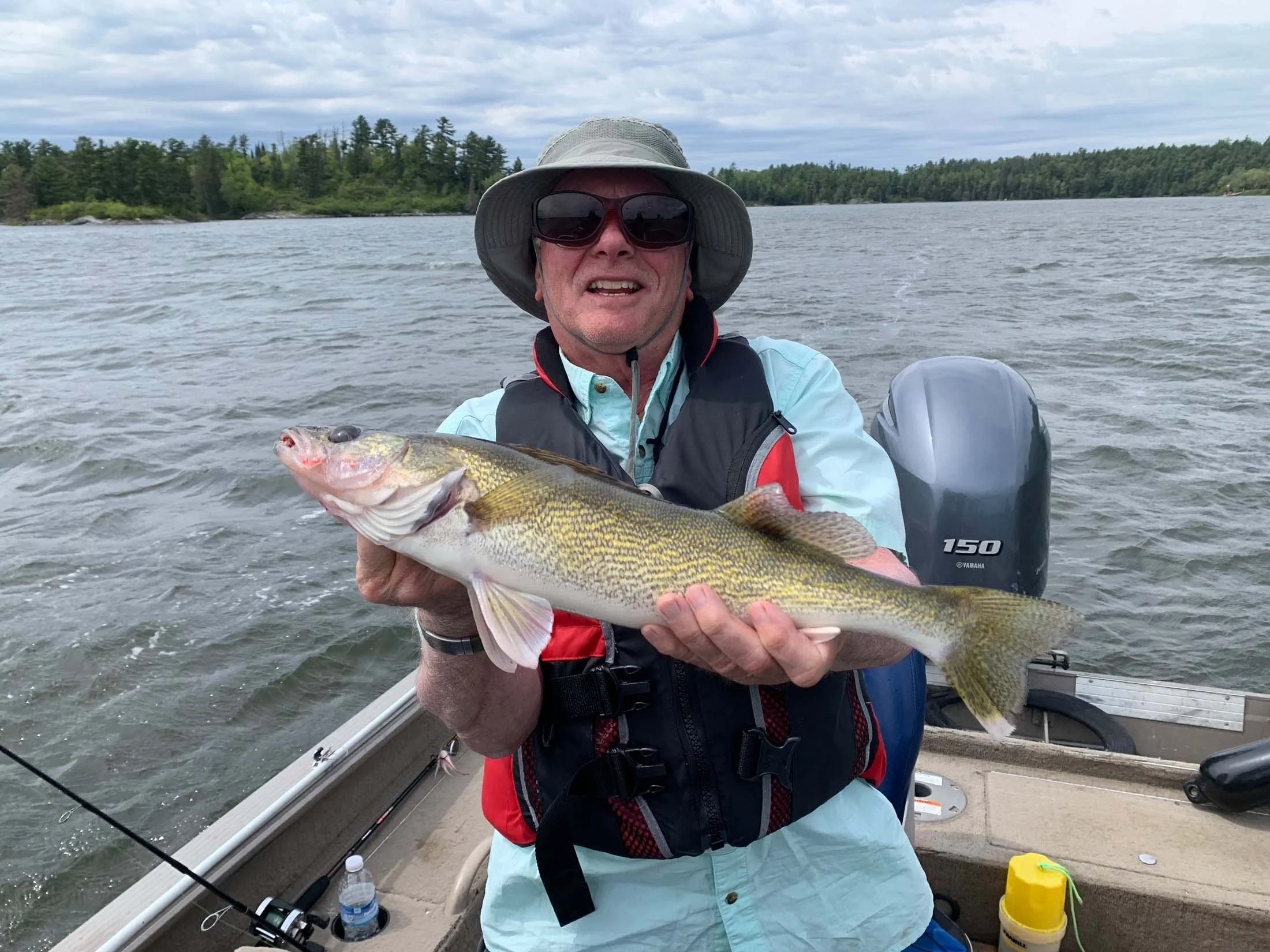 A man on a boat holding a large fish during a fishing trip on a lake with forested shoreline in the background.