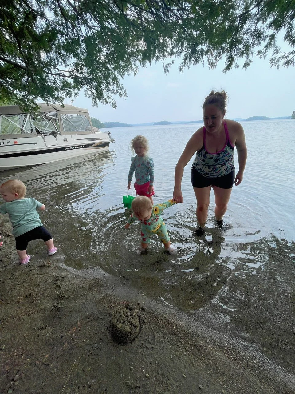 A woman with three young children at the edge of a lake, wading in the water under a tree overcast sky with a boat in the background.
