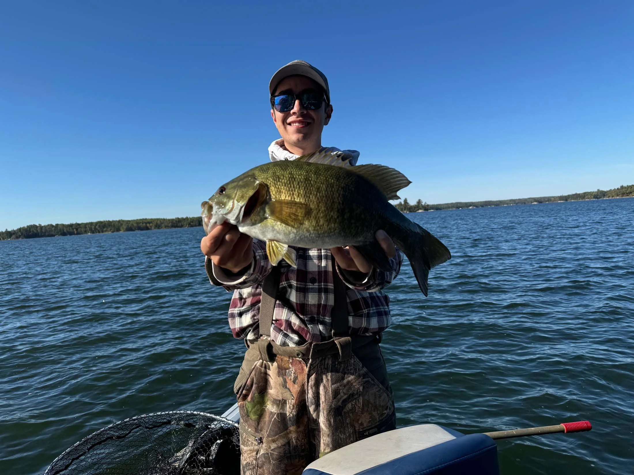 A man wearing a cap and sunglasses holding a large fish on a boat in a lake with a forested shoreline in the background under a clear blue sky.