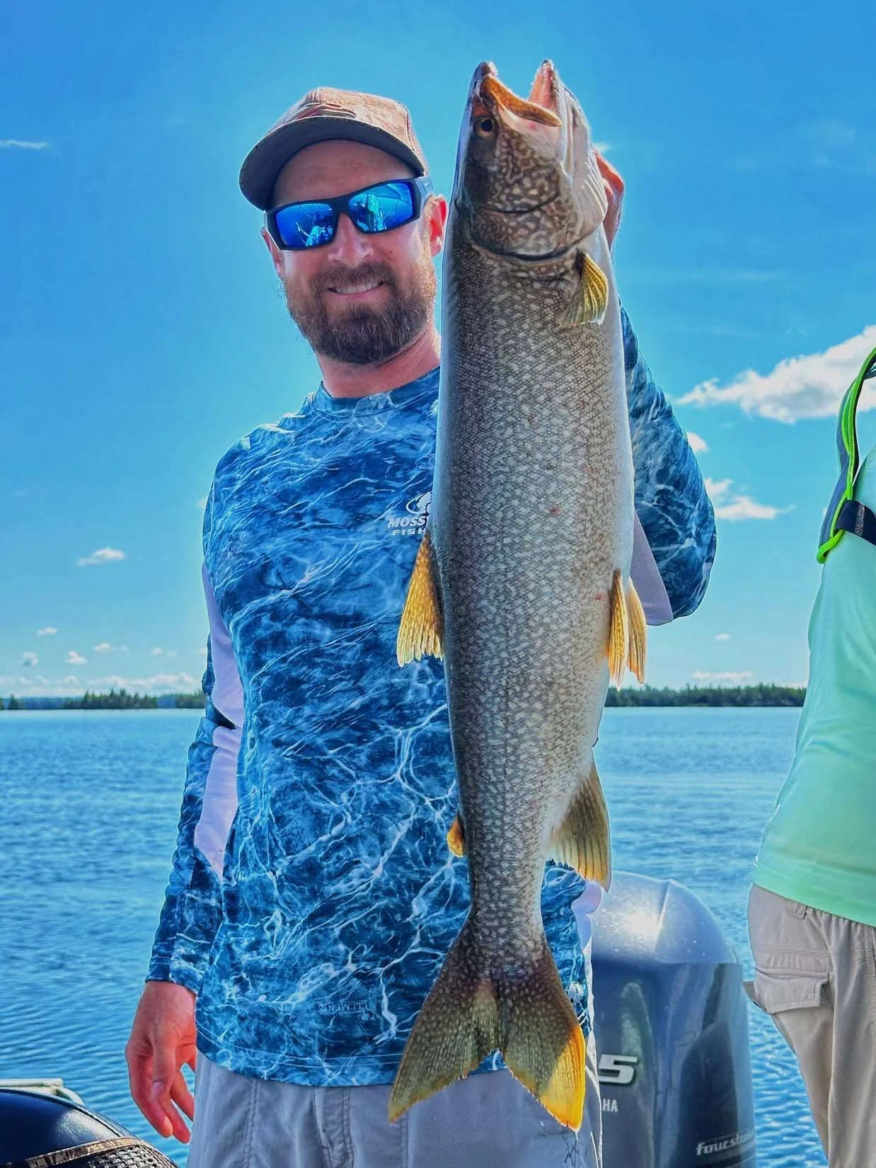 A man wearing a camouflage cap, blue sunglasses, and a blue patterned fishing shirt stands outside on a boat, holding a large fish with a long body, gill covers, and a wide open mouth, against a background of a body of water and a blue sky with some 