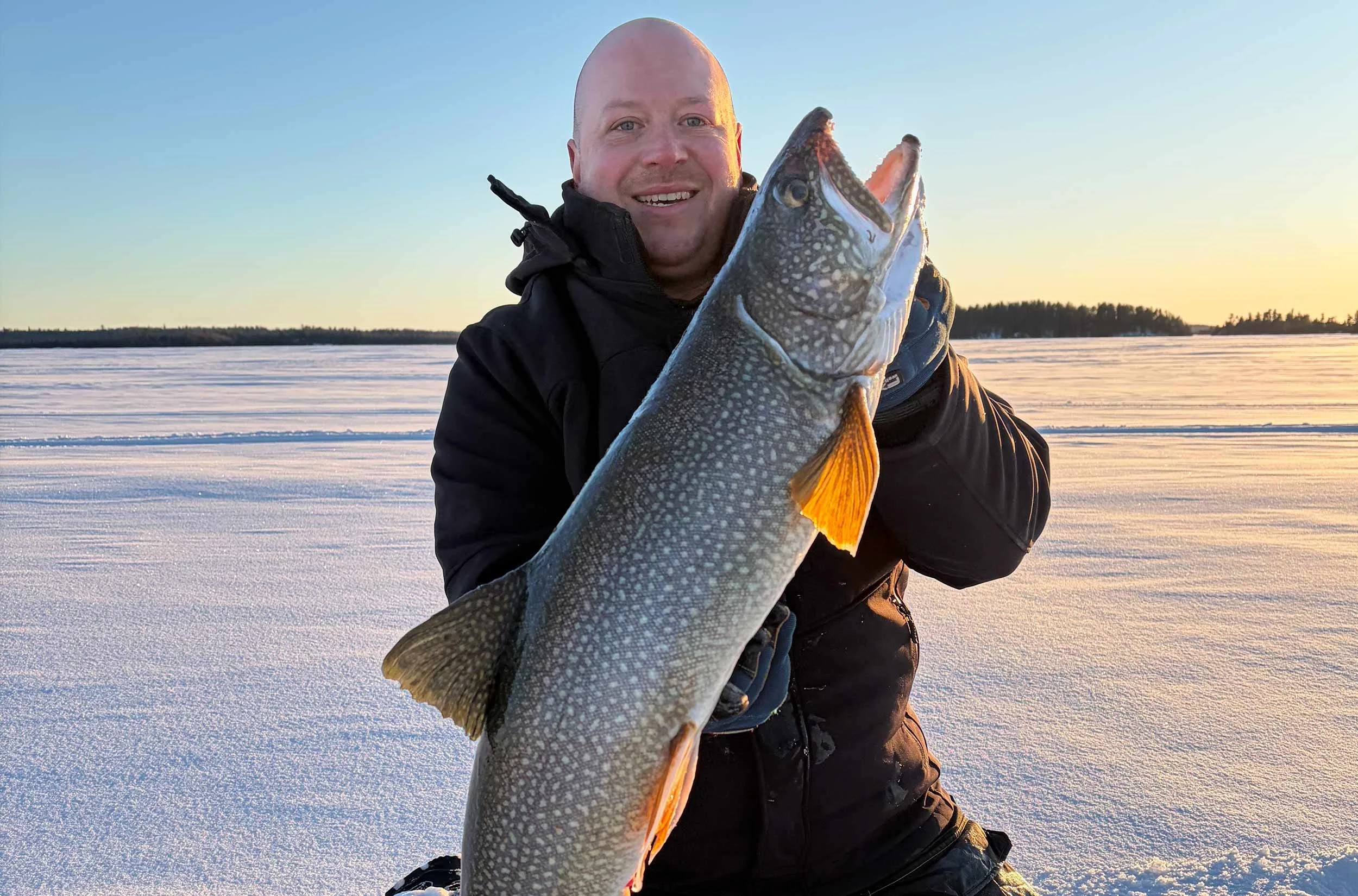 A man dressed in black winter clothing kneels on snow-covered ground while holding a large fish with light-colored, speckled scales and orange fins, during sunset with a distant forest silhouette.