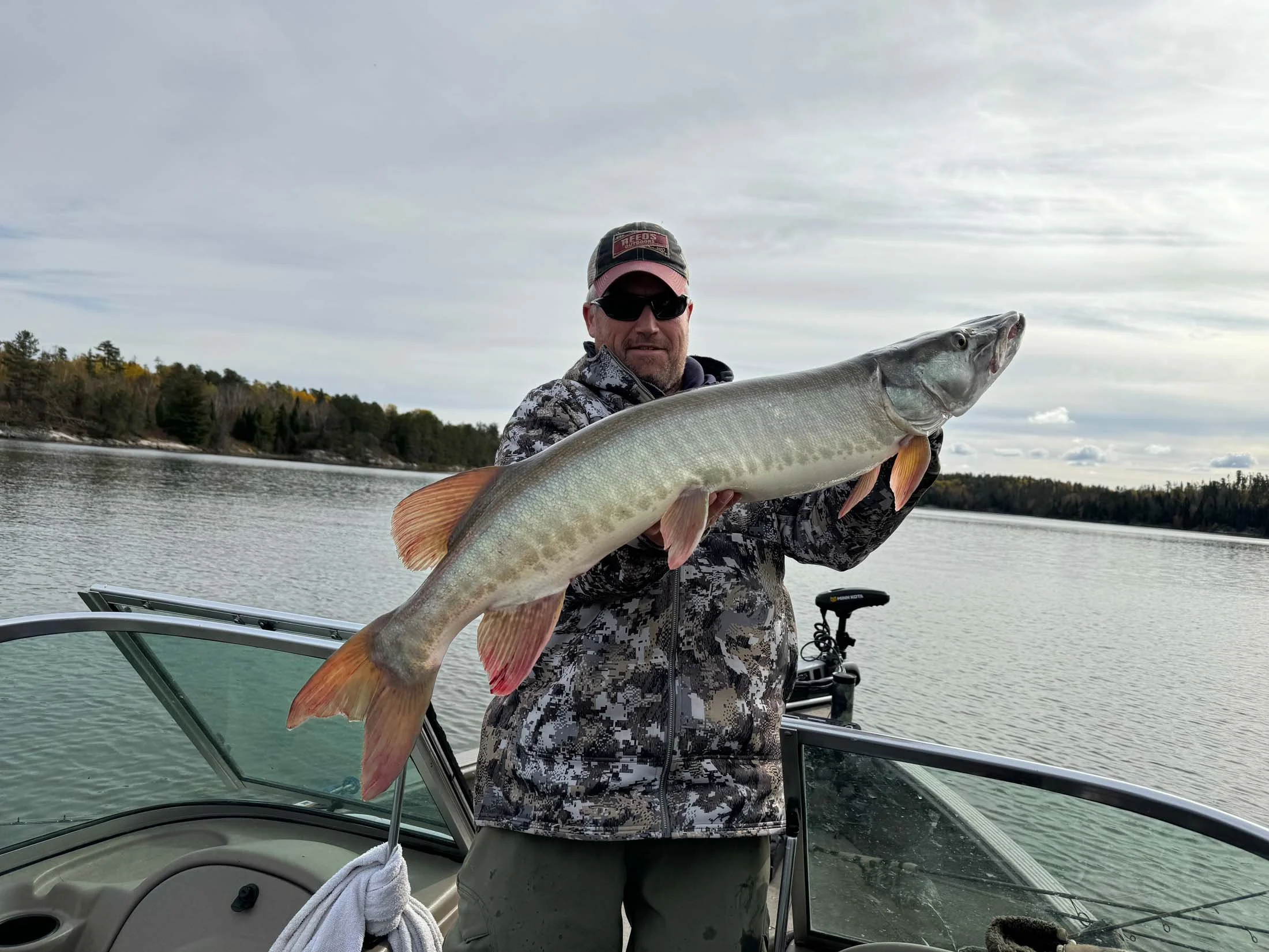 Man holding a large fish on a boat, with water and trees in the background.