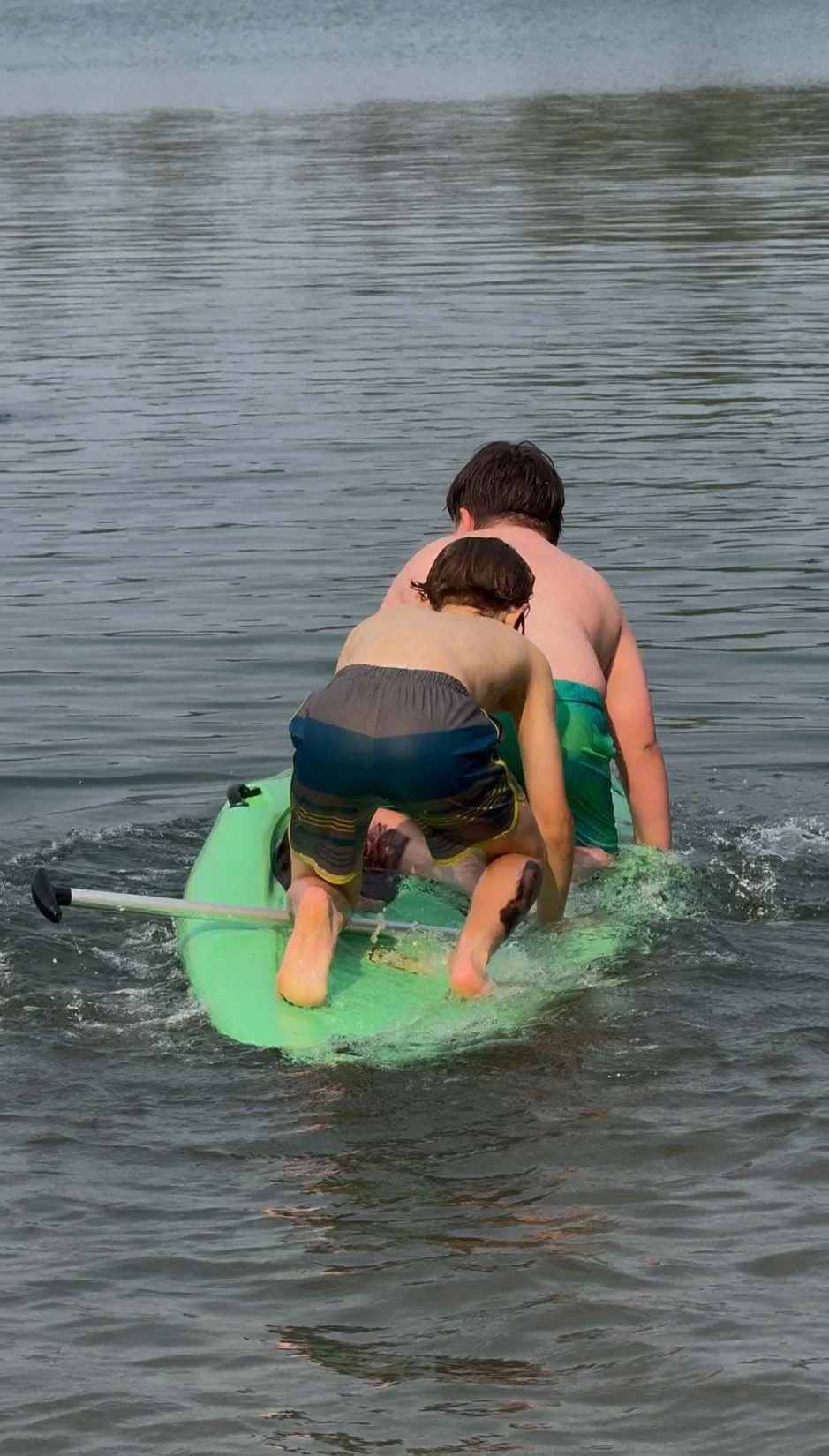 Two boys paddle a green kayak on a calm lake, with one sitting in front and the other behind, both wearing swim trunks.