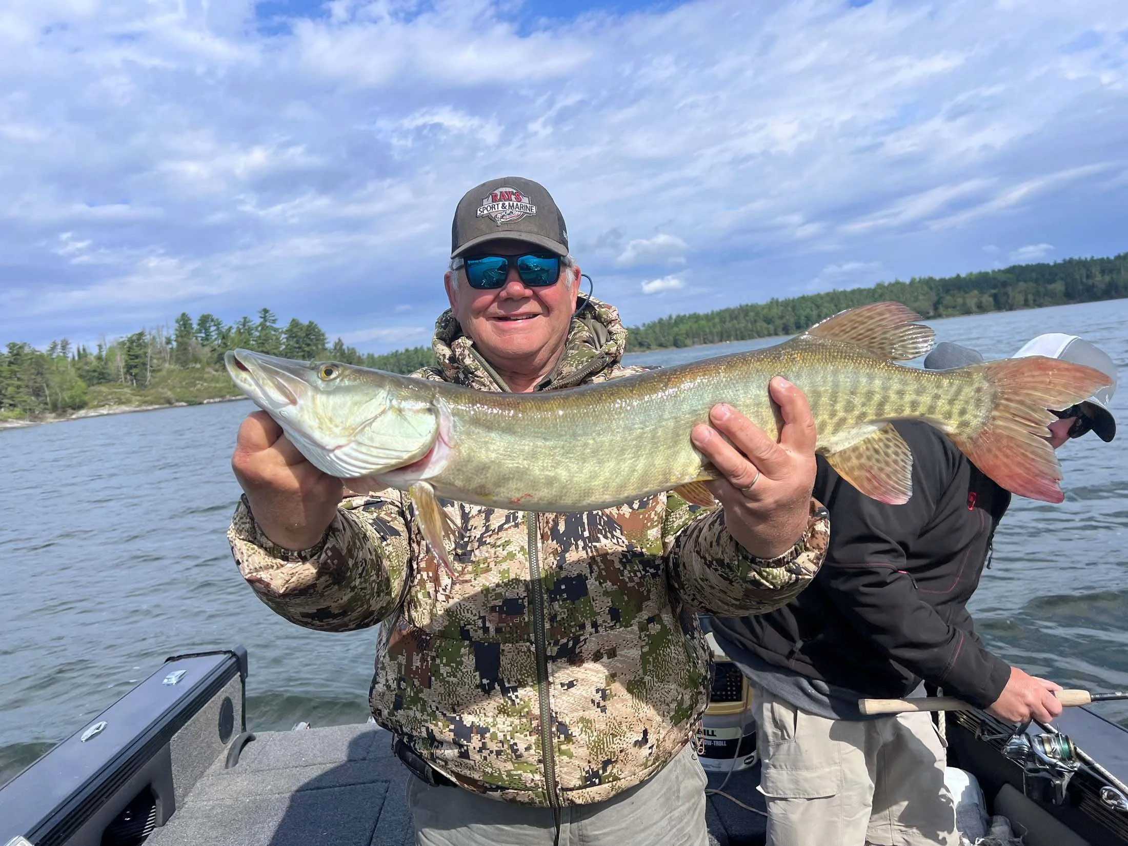 A man in camouflage clothing, wearing sunglasses and a cap, is smiling and holding a large fish he caught while on a boat on a lake.