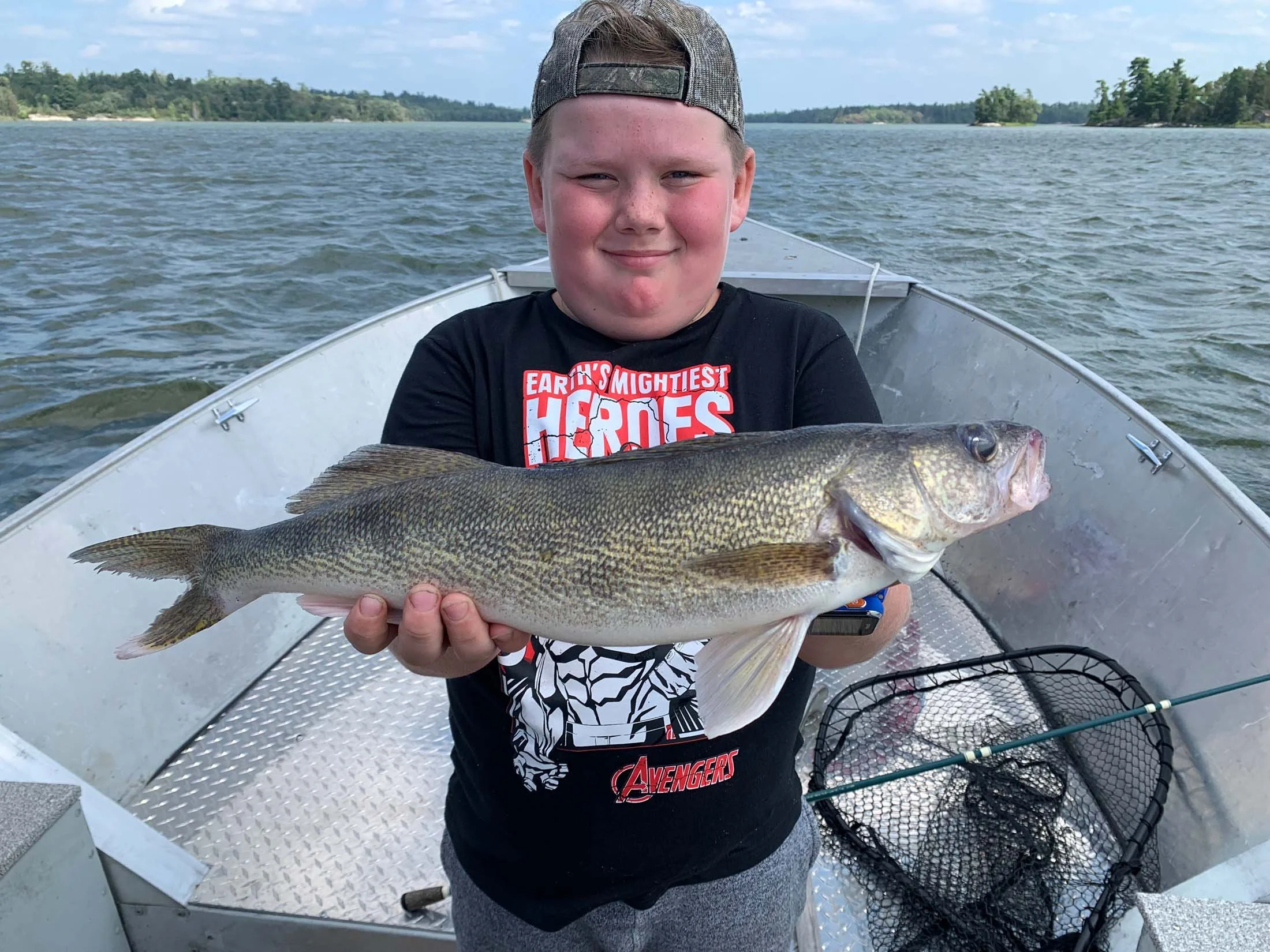 Young boy on a boat holding a large fish, with water and trees in the background, wearing a cap and an Avengers T-shirt.
