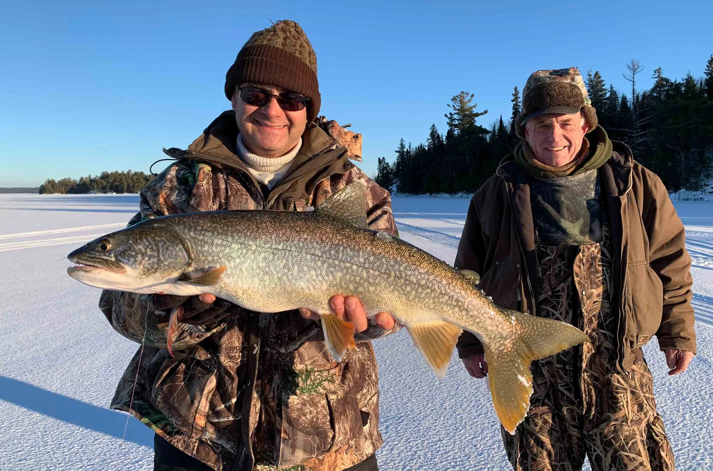 Two men dressed in winter camouflage holding a large fish, standing on a snowy landscape with a forested area in the background.