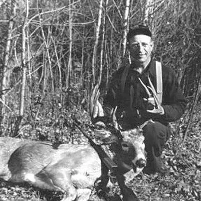 A man with glasses and a cap kneeling next to a large deer he has hunted, in a wooded outdoor setting.