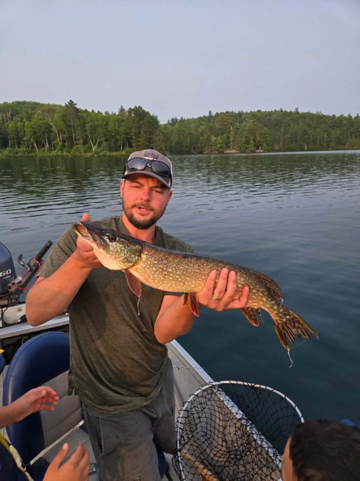 Man in boat holding a large fish, with trees and water in the background.
