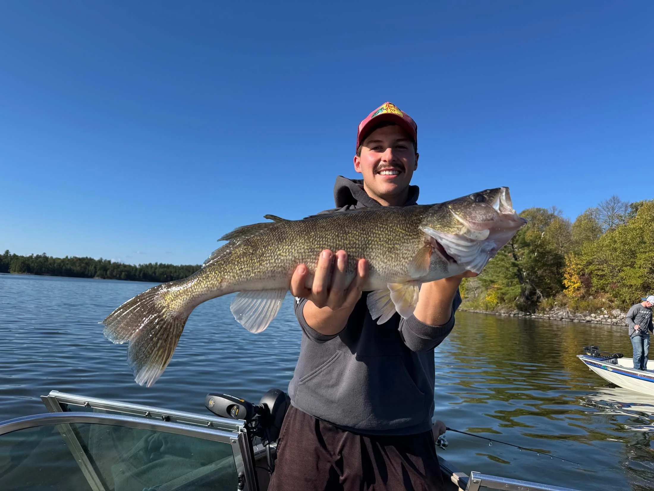 A young man wearing a red cap and a gray hoodie is smiling while holding a large fish with dark green and yellow scales. He is on a boat in a lake with trees and blue sky in the background, and another person is fishing nearby.
