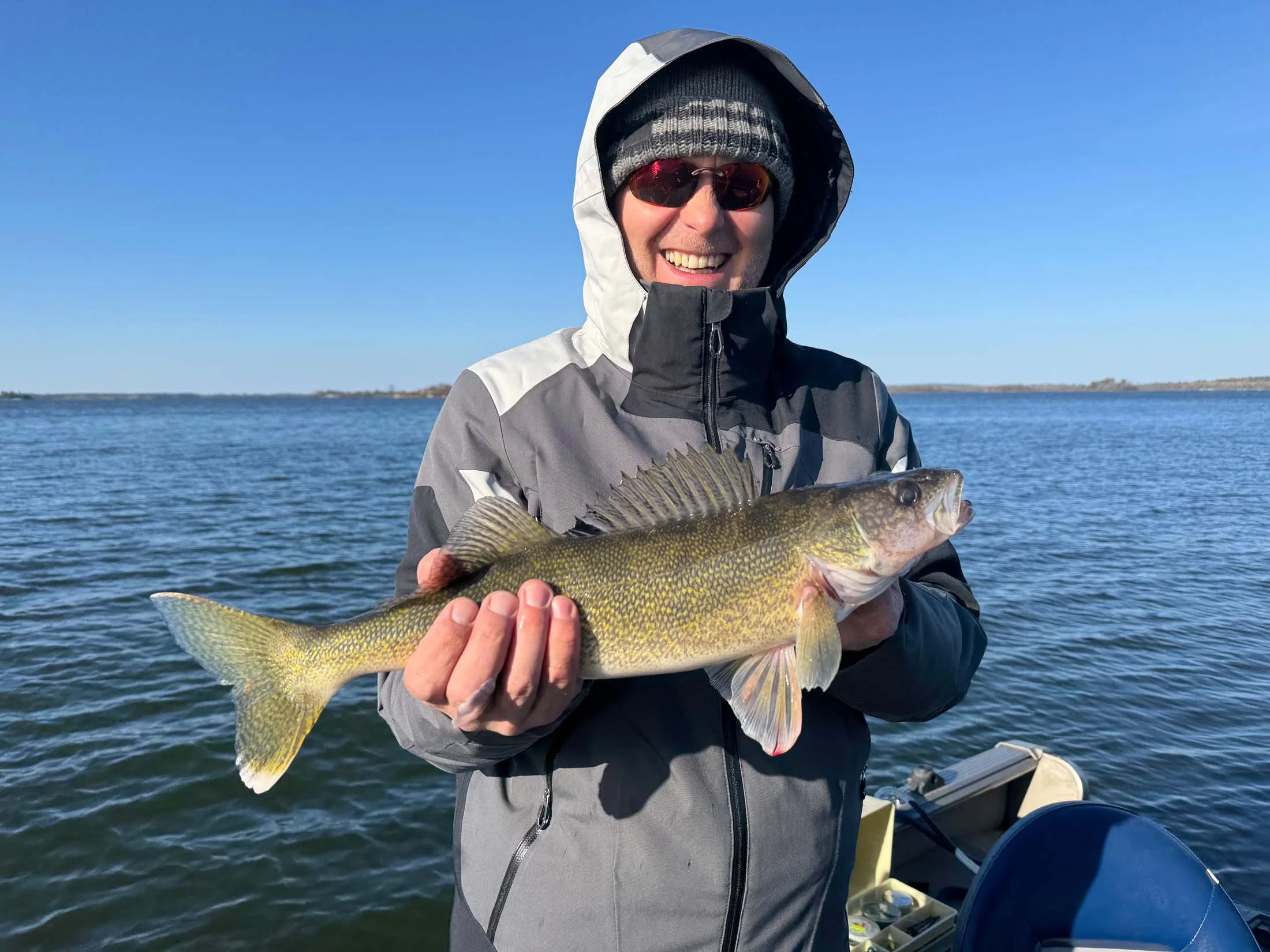 A man wearing a gray jacket, black and gray beanie, and sunglasses is smiling and holding a large fish with yellow and brownish-green scales while standing on a boat on a lake with a clear blue sky in the background.