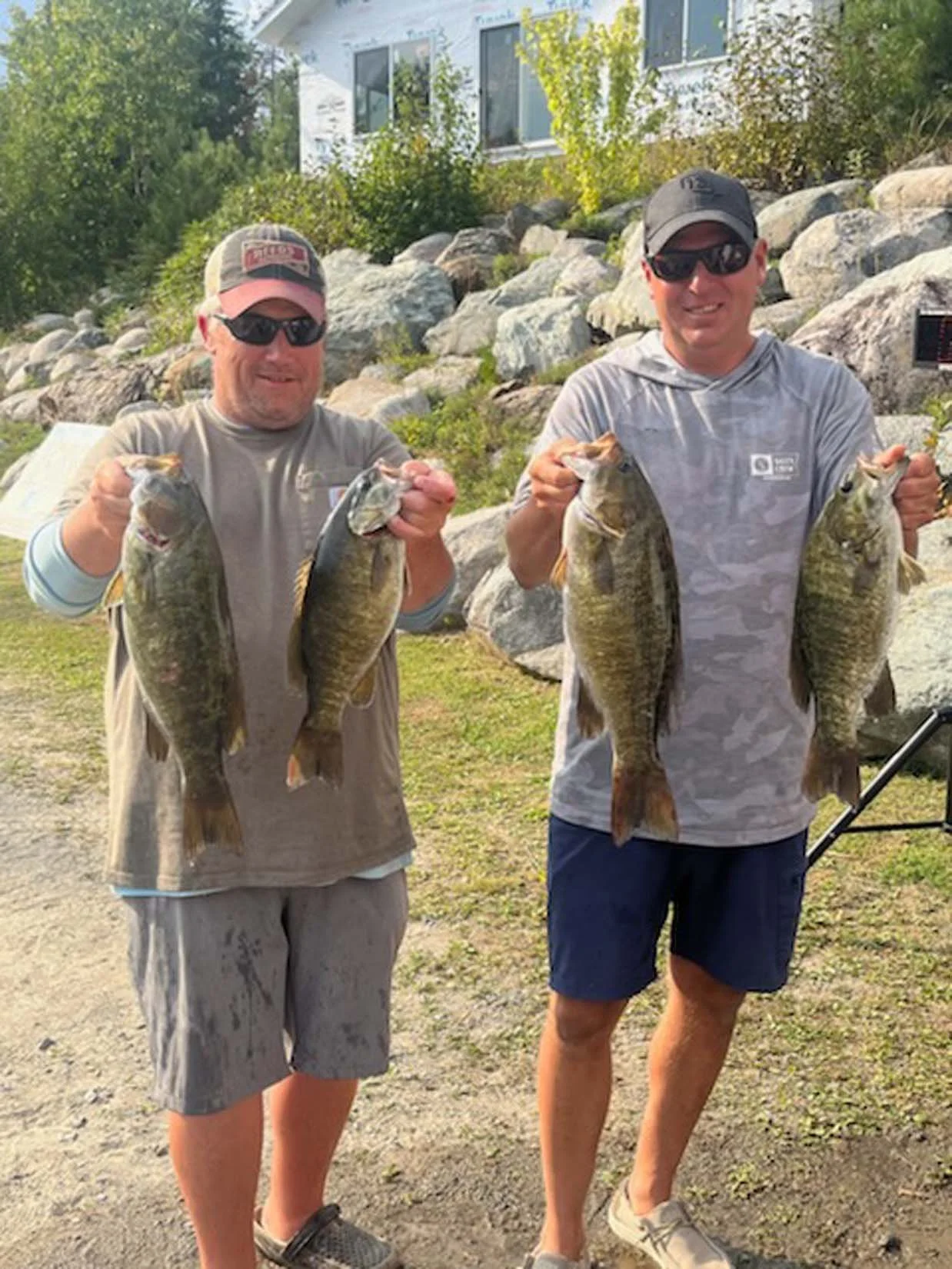 Two men standing outdoors holding four large fish, smiling, wearing sunglasses and casual clothing, with rocks, trees, and a building in the background.