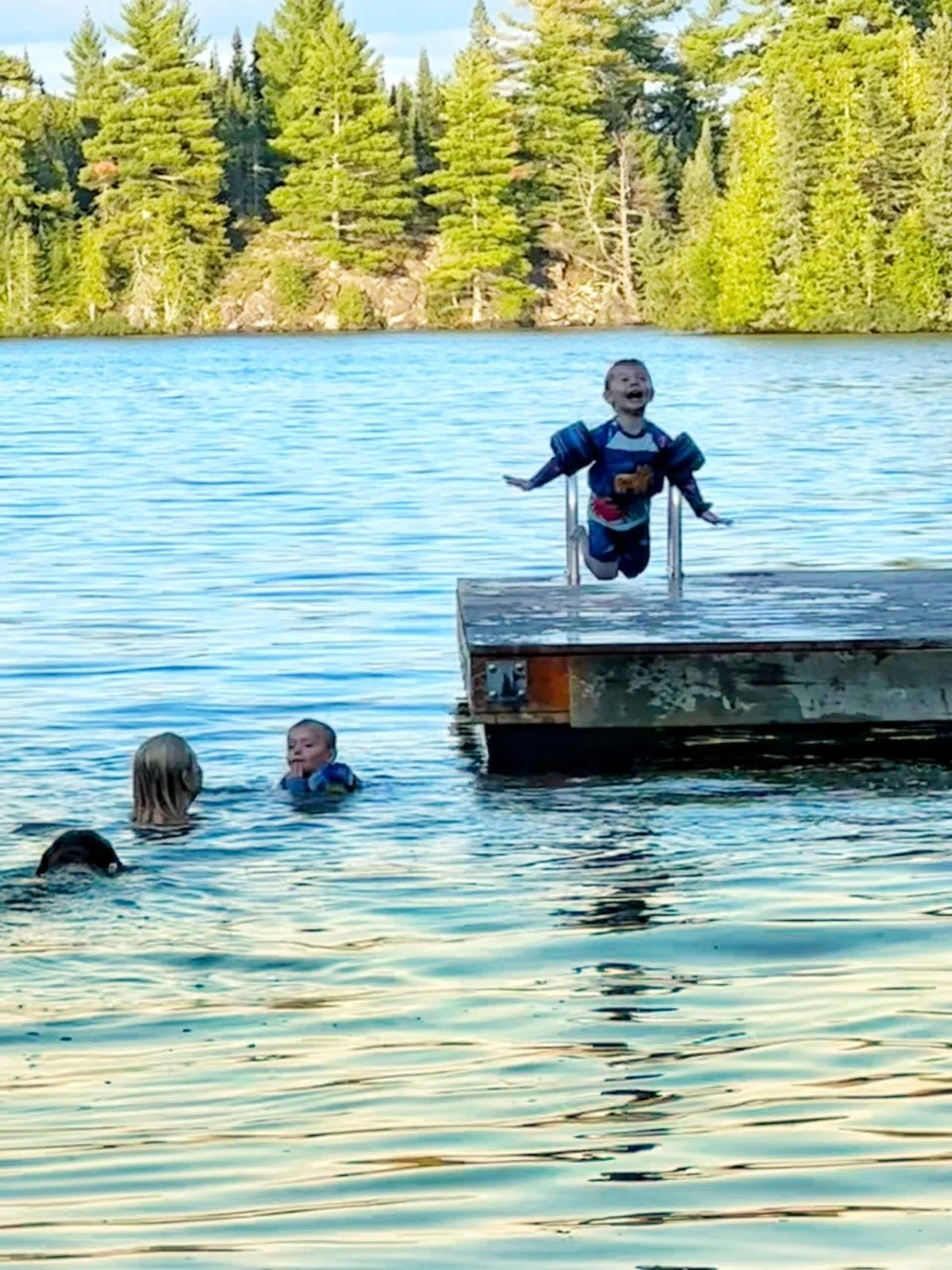 Children swimming and playing near a dock on a lake with a forested shoreline and trees in the background.