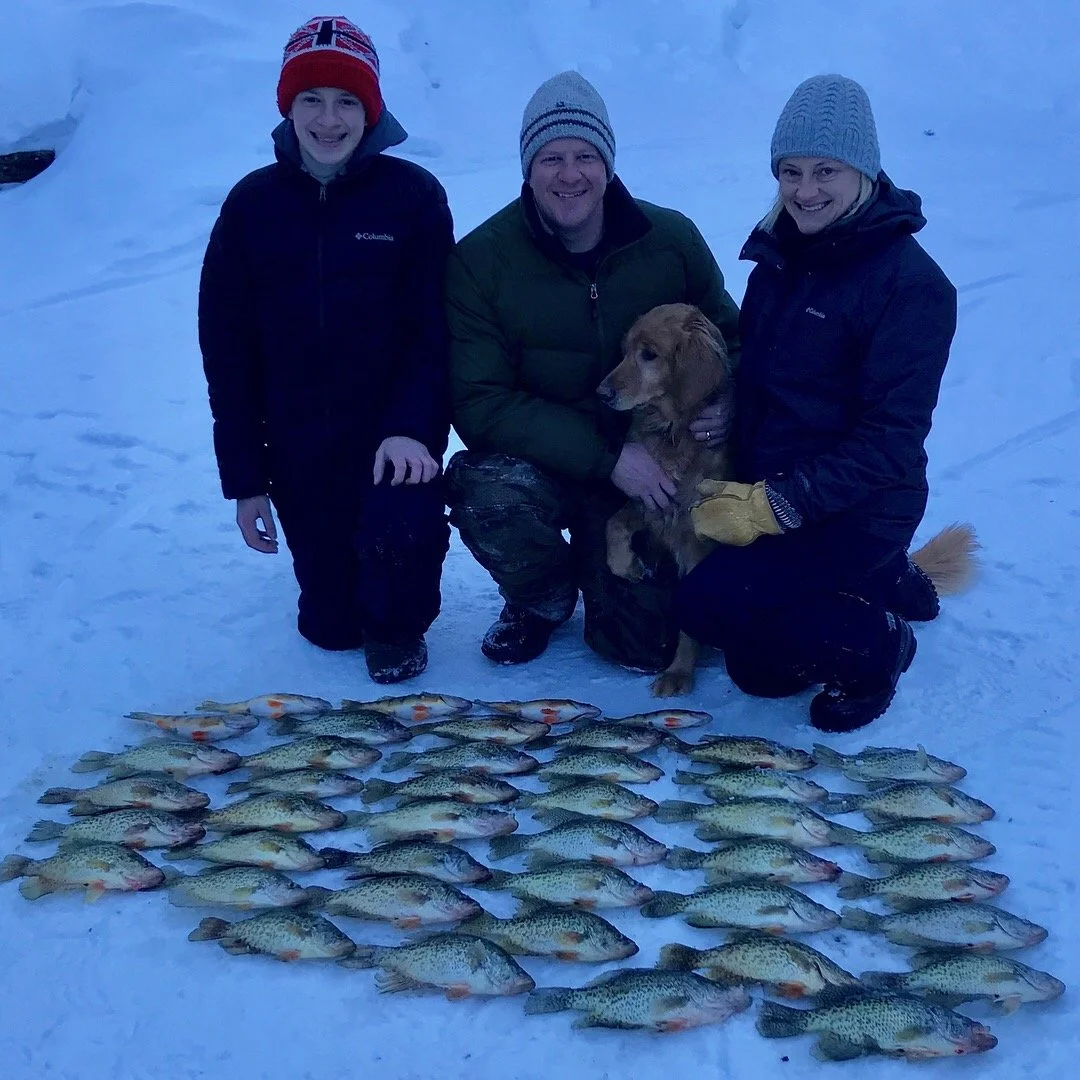 A family of three with a dog kneeling on snow in winter outdoors, displaying their catch of numerous fish laid out on the snow. The family members are dressed in winter clothing, smiling, with a young boy on the left, a man in the middle, and a woman