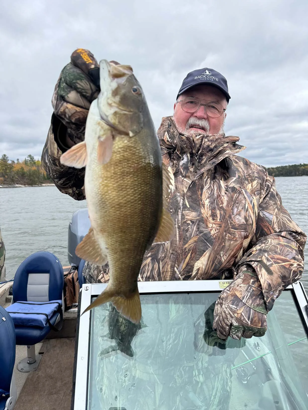 An older man in camouflage jacket and cap holding a largemouth bass fish on a boat on a lake with a cloudy sky in the background.