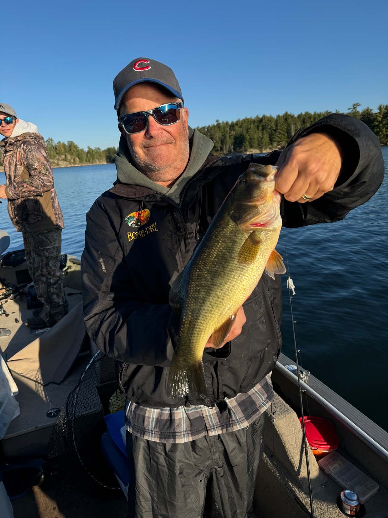 A man holding a large fish on a boat on the water, with another person in the background, trees and a clear blue sky.