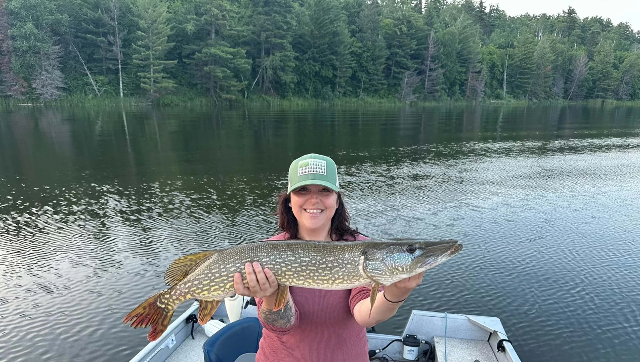 A woman smiling on a boat holding a large fish with a lake and forest in the background.