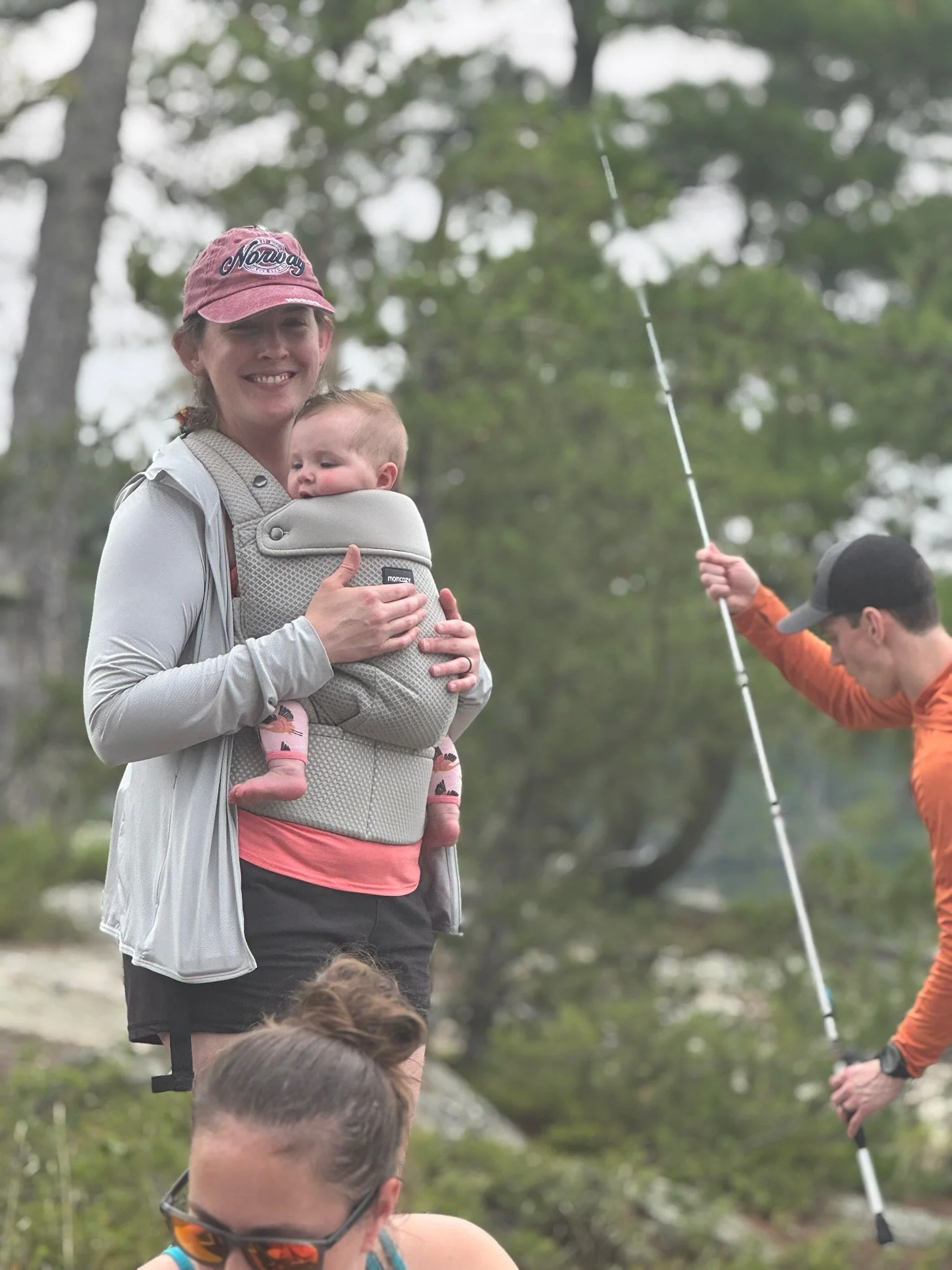 A woman smiling and carrying a baby in a front baby carrier outdoors, with three other people holding fishing rods in a wooded area.