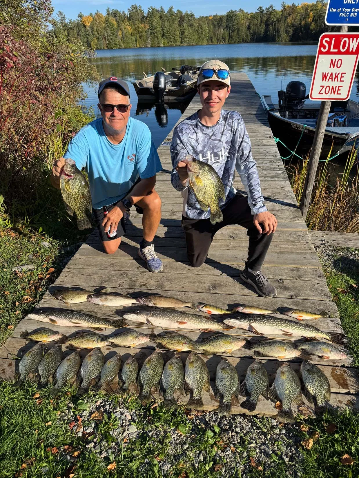 Two men kneeling on a wooden dock holding fish, with multiple fish laid out in front of them. Behind them, there is a lake with boats docked and a forested shoreline. A red and white sign reads 'Slow No Wake Zone'.