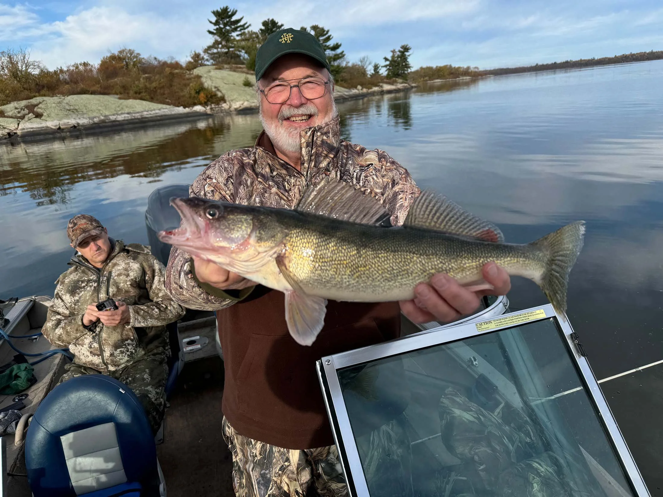 A man in camouflage clothing holding a large fish on a boat, with calm water and trees in the background, and another person sitting near him wearing camo, with fishing equipment on the boat.