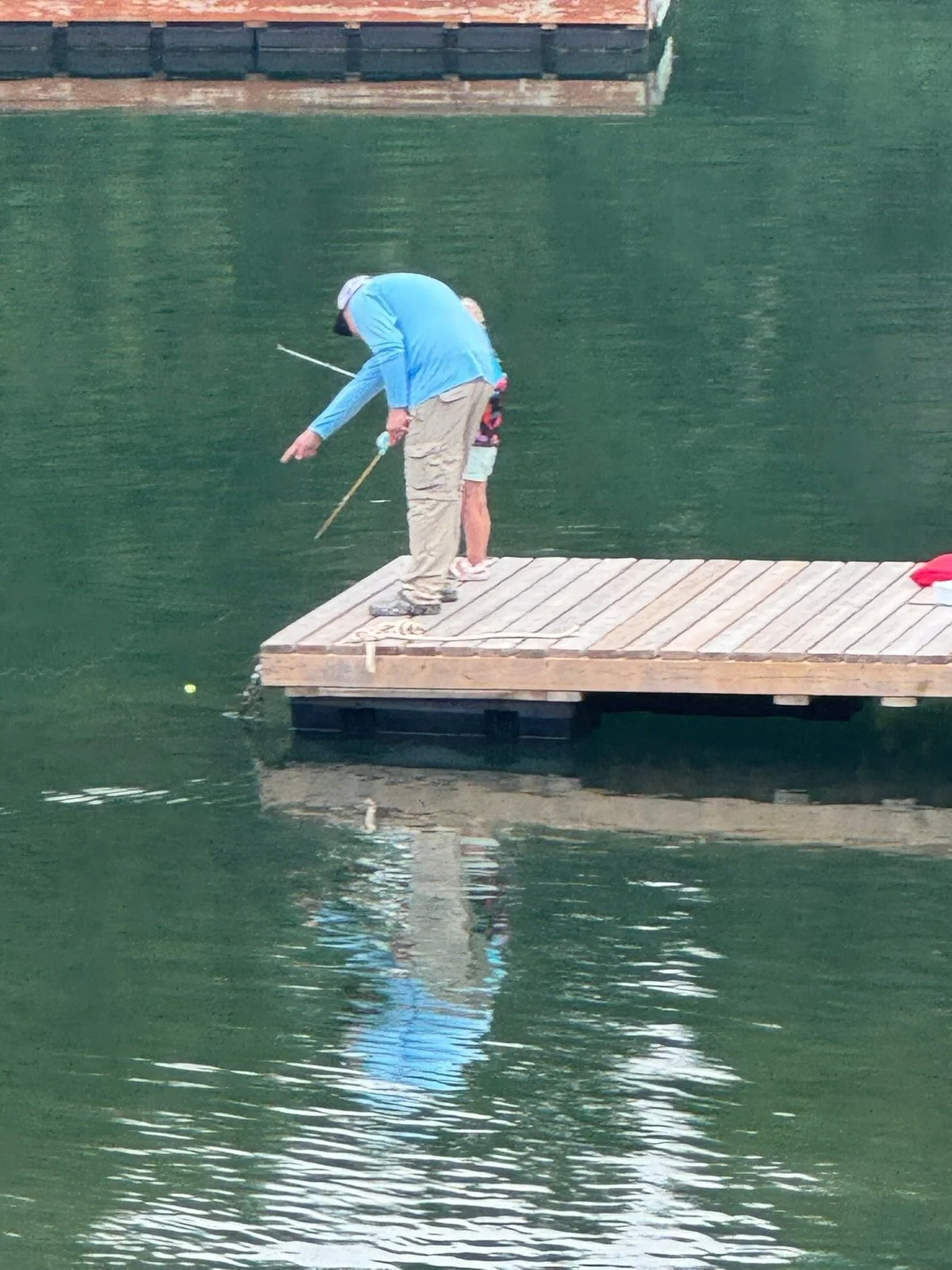 A person in a blue jacket standing on a wooden dock fishing in a green body of water, with a child standing nearby.