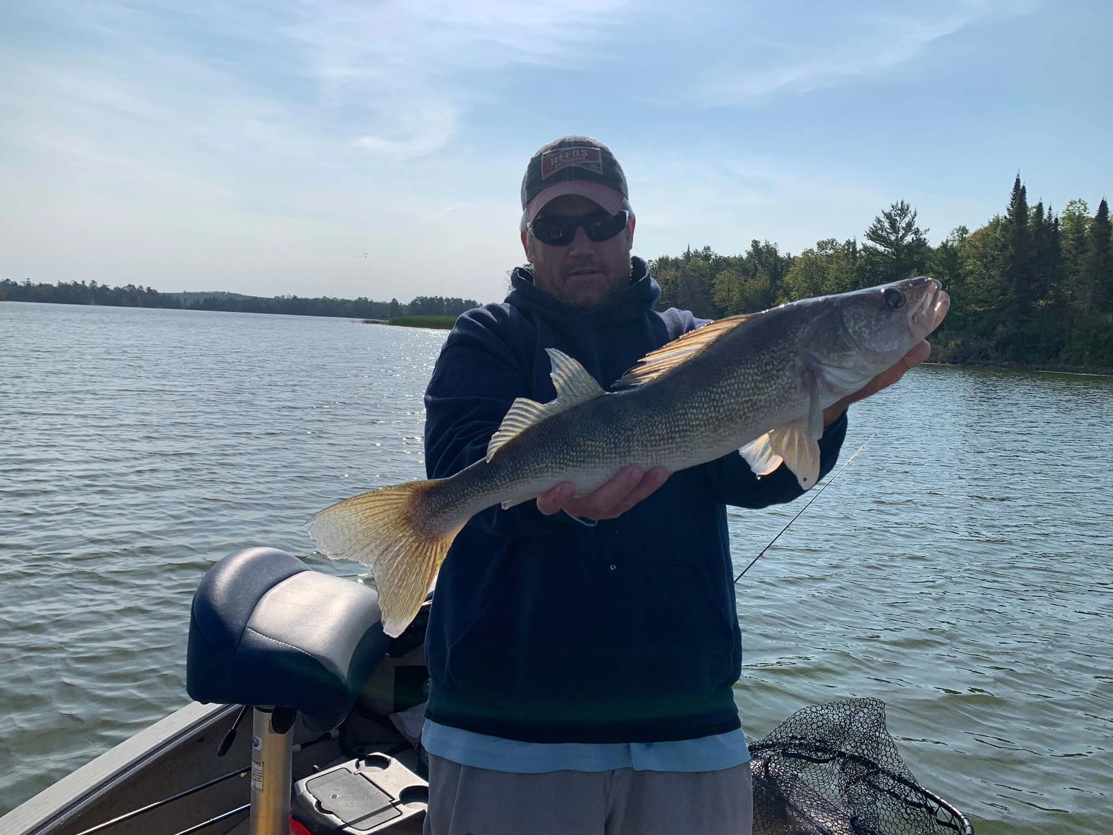 Man wearing sunglasses, a baseball cap, and a hoodie on a boat, holding a large fish over water with trees in the background.