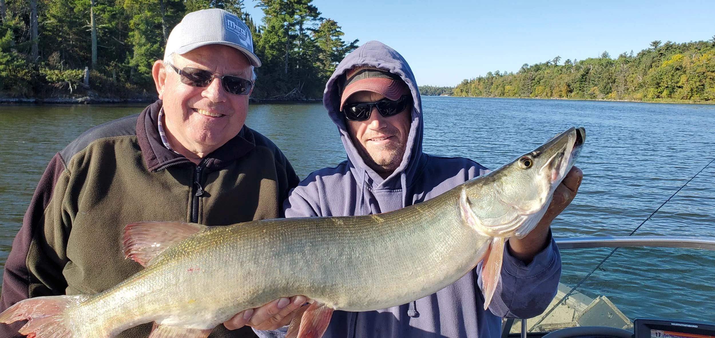 Two men on a boat holding a large fish with a river and forest in the background.