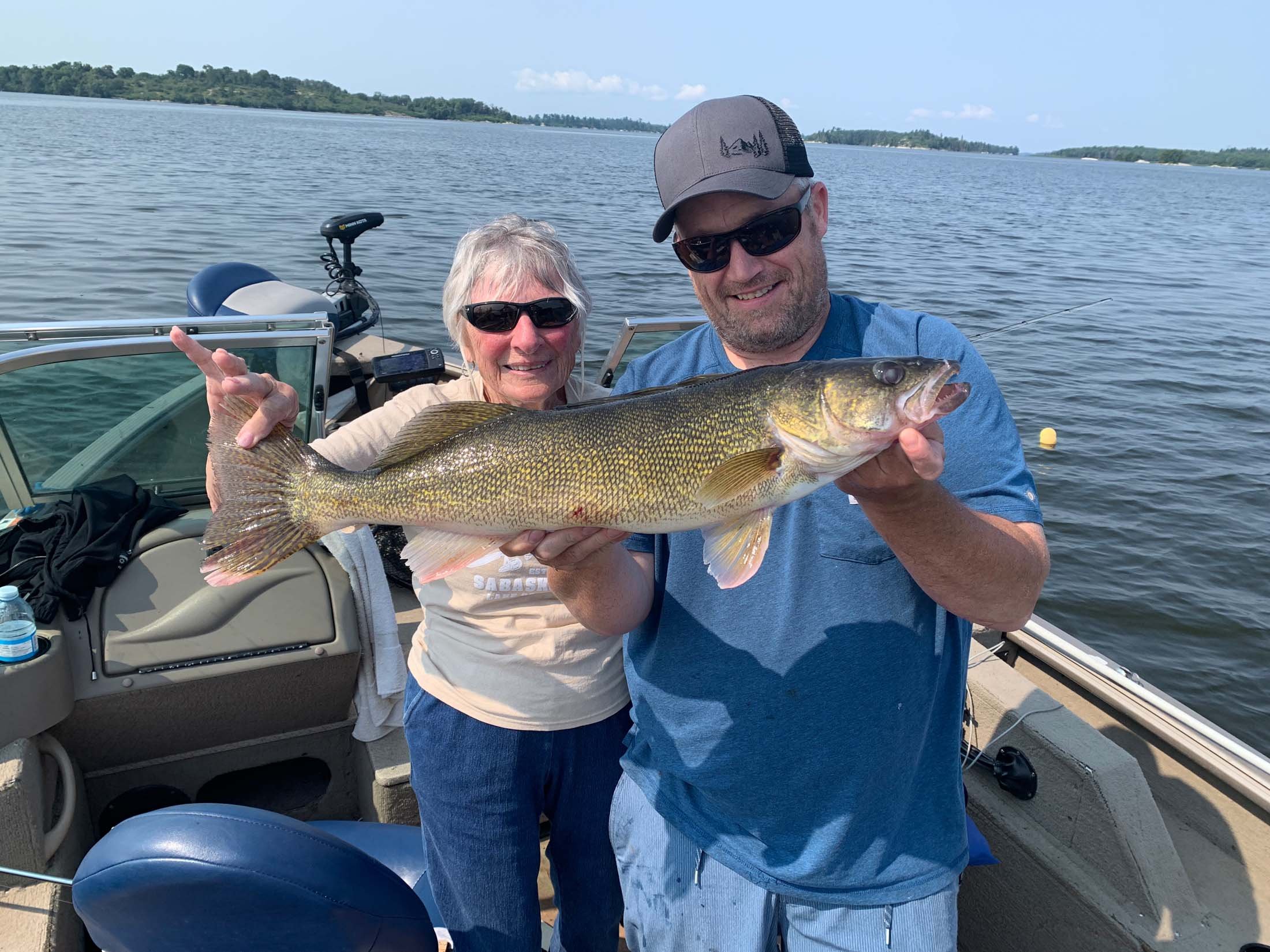 Two people on a boat holding a large fish, with a lake and distant land in the background. The woman on the left is wearing sunglasses and a beige shirt, the man on the right is wearing a gray cap, sunglasses, and a blue shirt. They are smiling and d