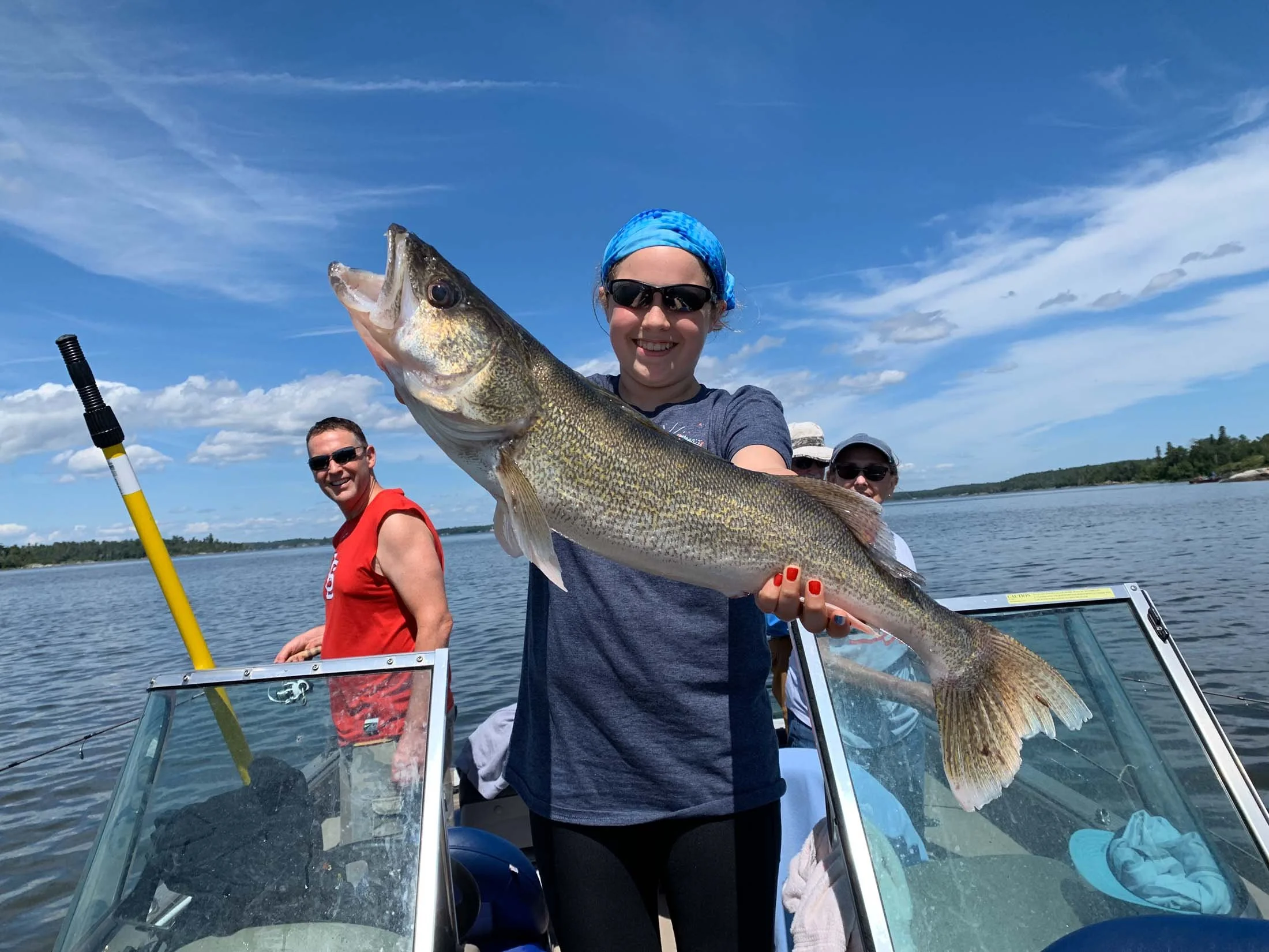 A woman in sunglasses and a blue headscarf holding a large fish on a boat, with two people in the background, water, and a clear sky.