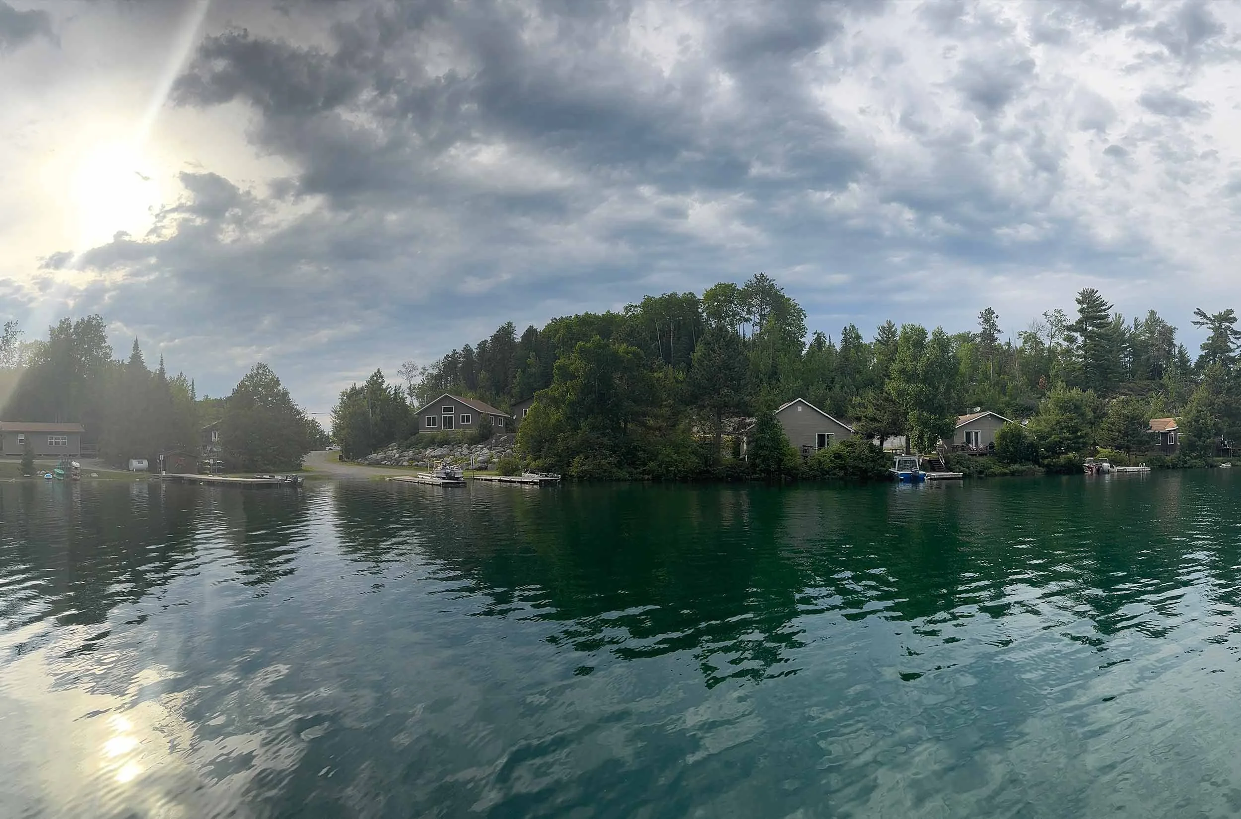 Calm water reflecting cloudy sky with some houses and trees along the shoreline, and a few boats docked at the water's edge.