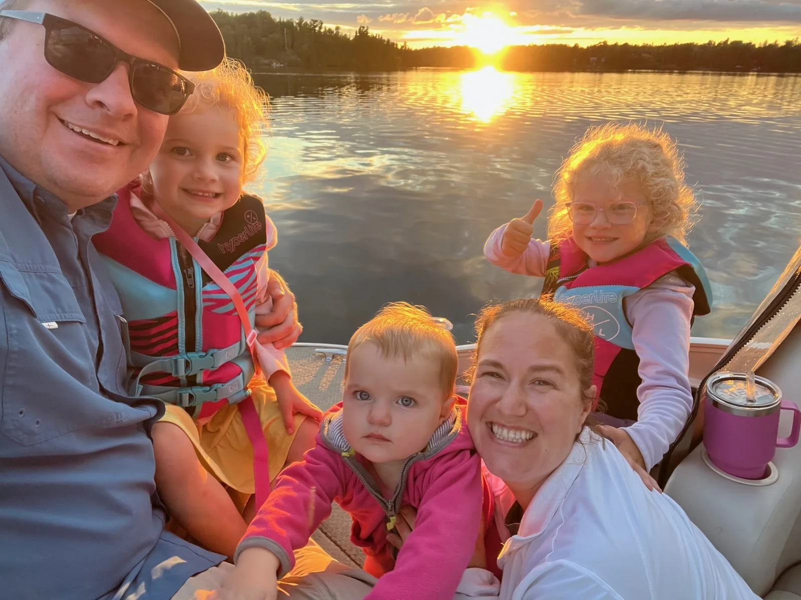 A family of six enjoying a boat ride on a lake during sunset, smiling and making gestures, with a scenic water and forest background.