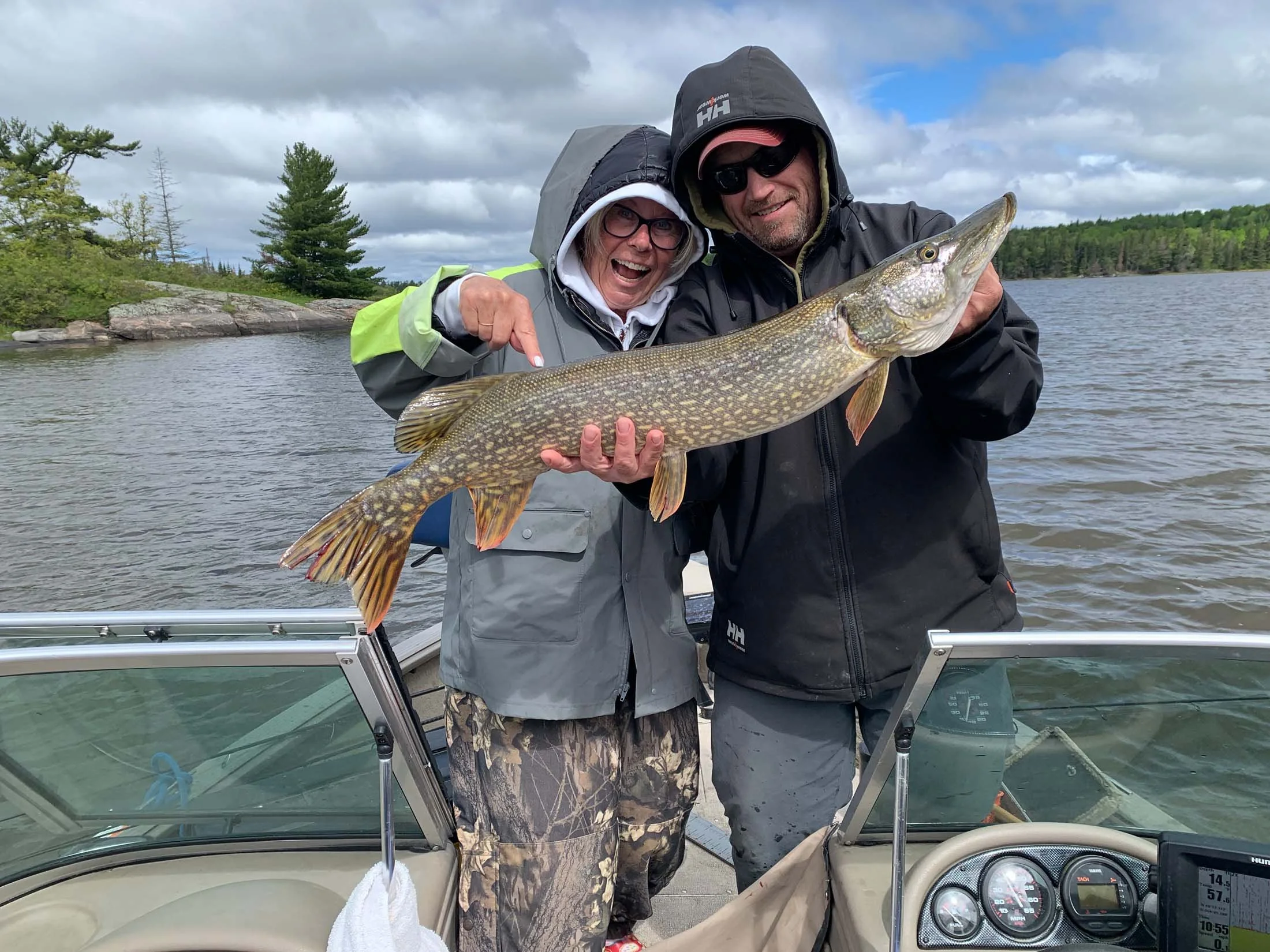 Two people on a boat holding a large fish, with trees and water in the background, wearing rain jackets and hats.