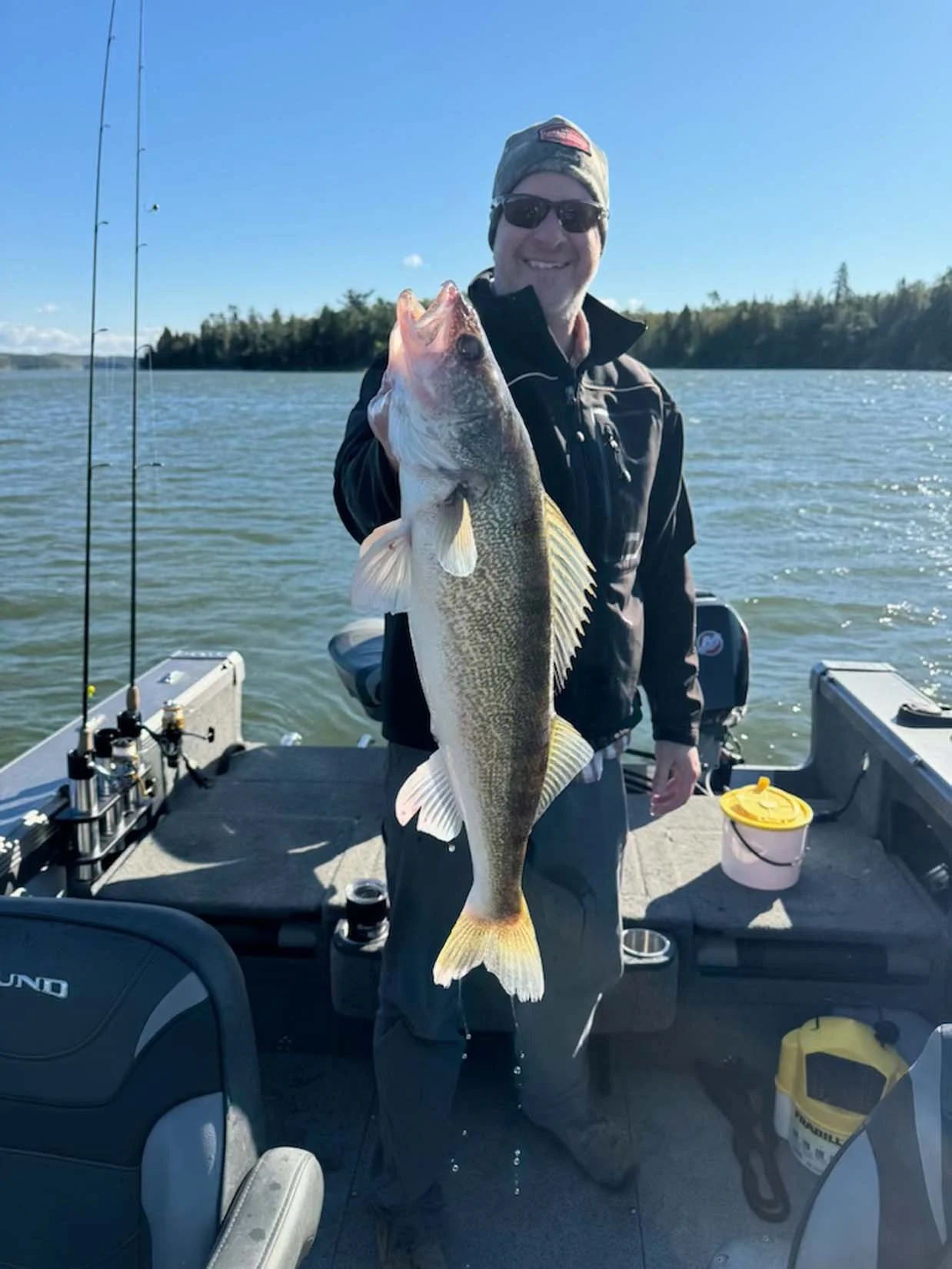Man on a boat holding a large fish with a body of water and trees in the background under a clear sky.