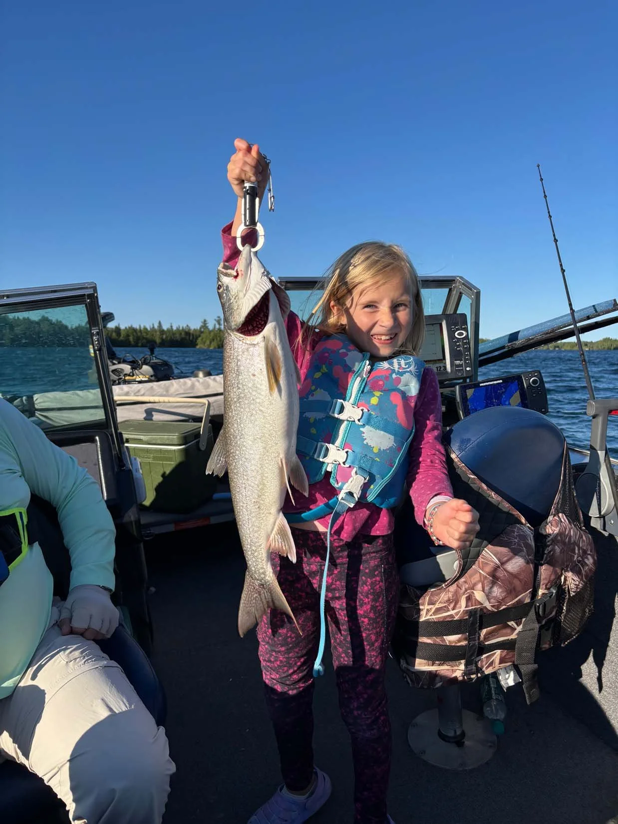 A young girl smiling and holding up a large fish she caught while on a boat. The boat is on a body of water, with a clear blue sky and distant trees in the background.