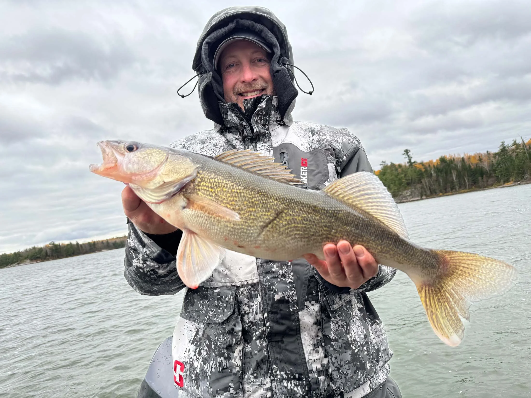 A smiling man wearing a camouflage jacket and a hood, holding a large fish over water during cloudy weather.