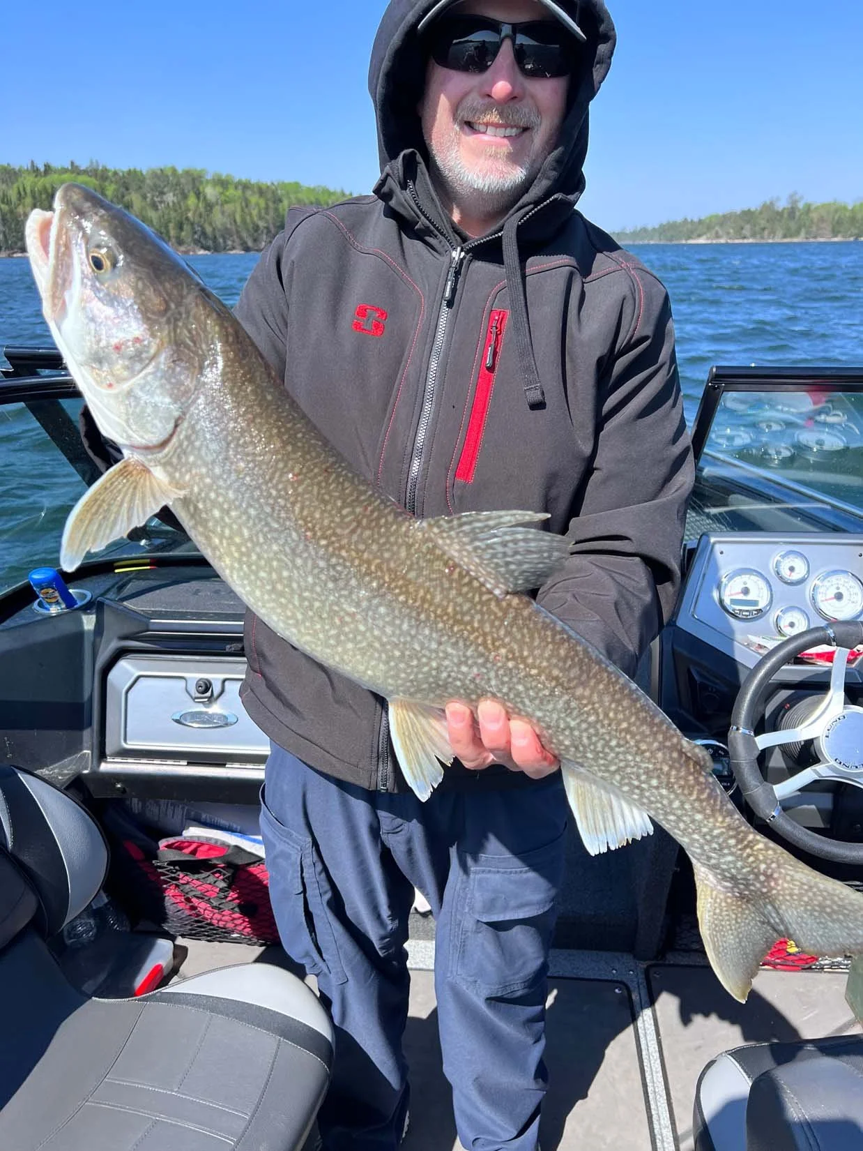 A man wearing a gray hoodie and sunglasses holding a large fish on a boat with water and trees in the background.
