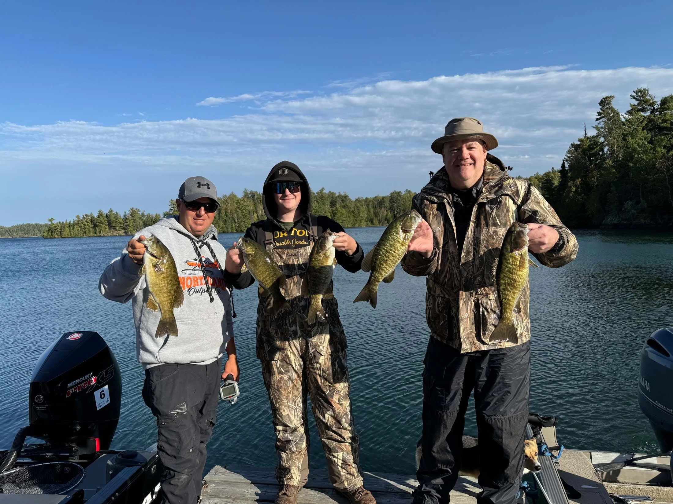Three men standing on a boat with a lake and trees in the background, each holding a fish they caught, during daytime with clear sky.